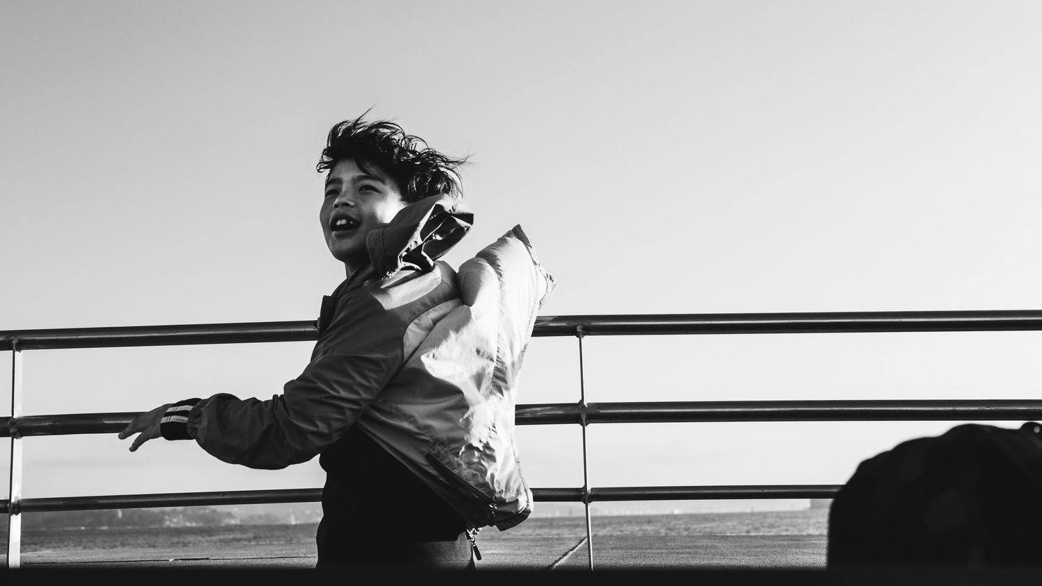 Black and white photo of young person leaning on metal railing outdoors against bright sky.