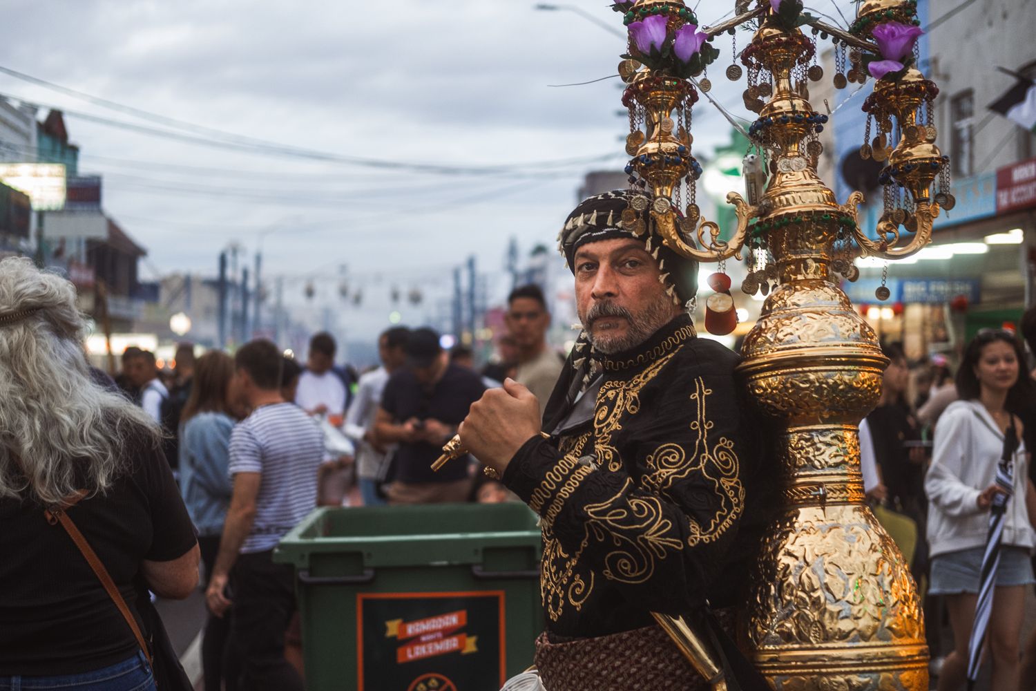 Man in ornate traditional costume holds decorative golden candelabras at a street festival.