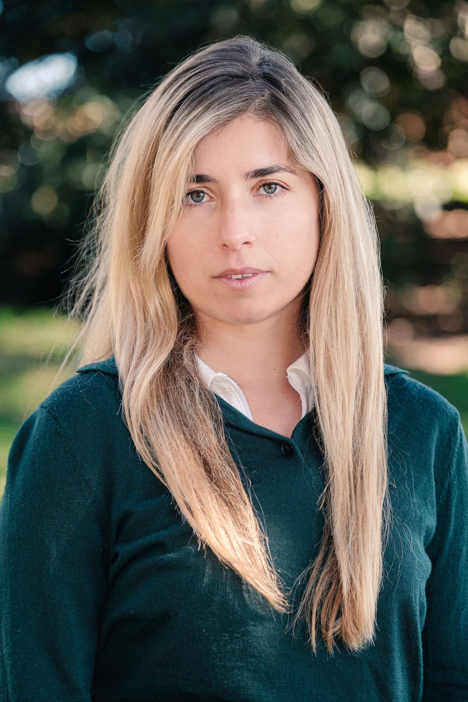Young blonde woman in green sweater poses outdoors with bokeh tree background in natural light portrait.