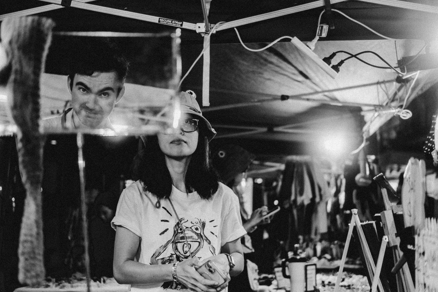 Young woman stands under market tent at night surrounded by hanging decorations in monochrome photo.