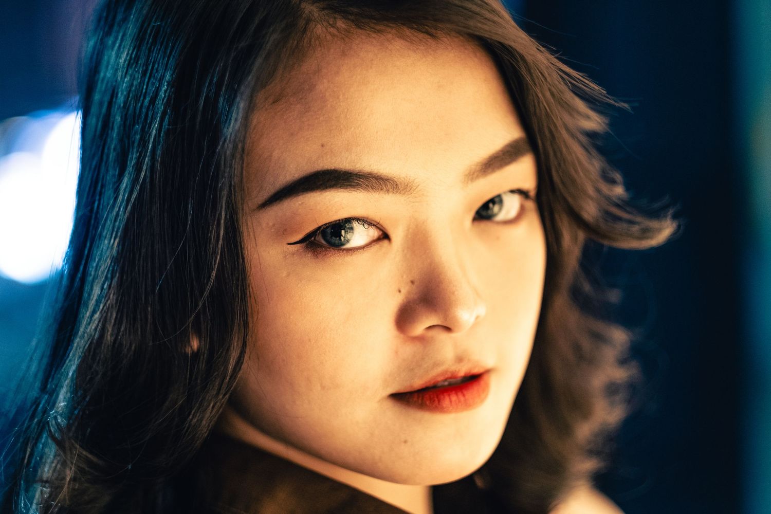 Close-up portrait of a young Asian woman with dark hair and red lips under blue and warm lighting.