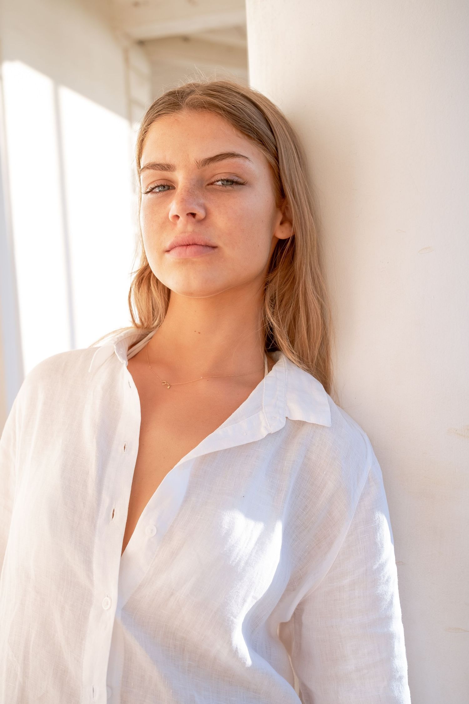 Young blonde woman in an open white linen shirt leaning against a white wall in sunlight.