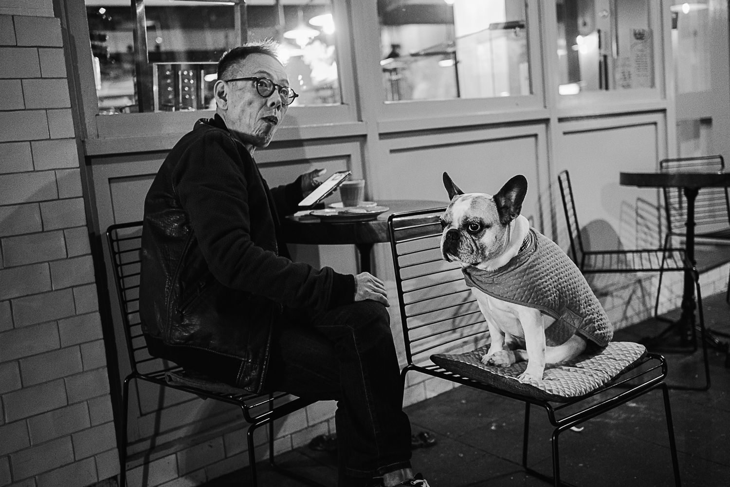 An elderly man reads at an outdoor cafe table while a French Bulldog in a sweater sits attentively beside him.