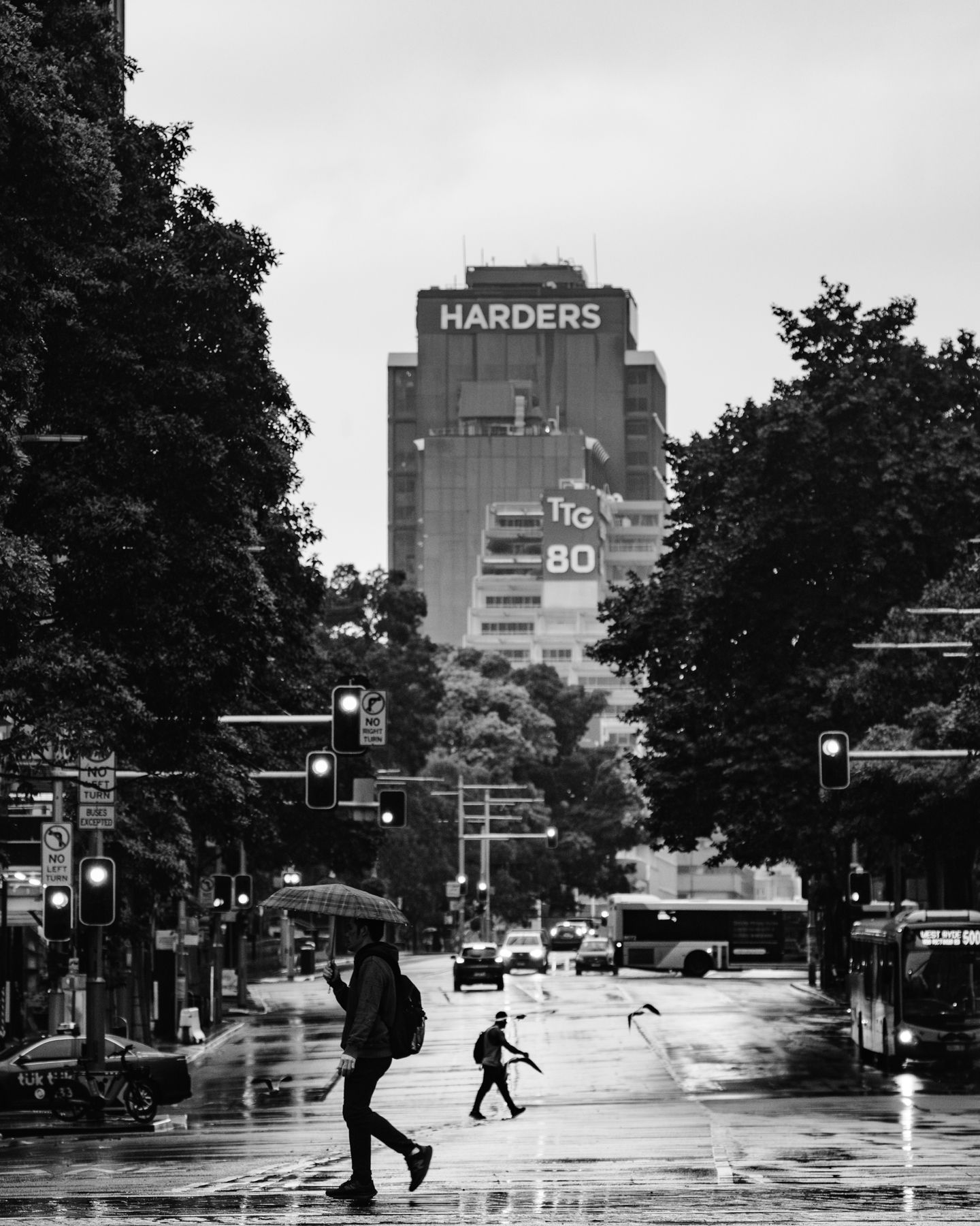 Pedestrians cross a rain-soaked city street in black and white with a Harders building in the background.