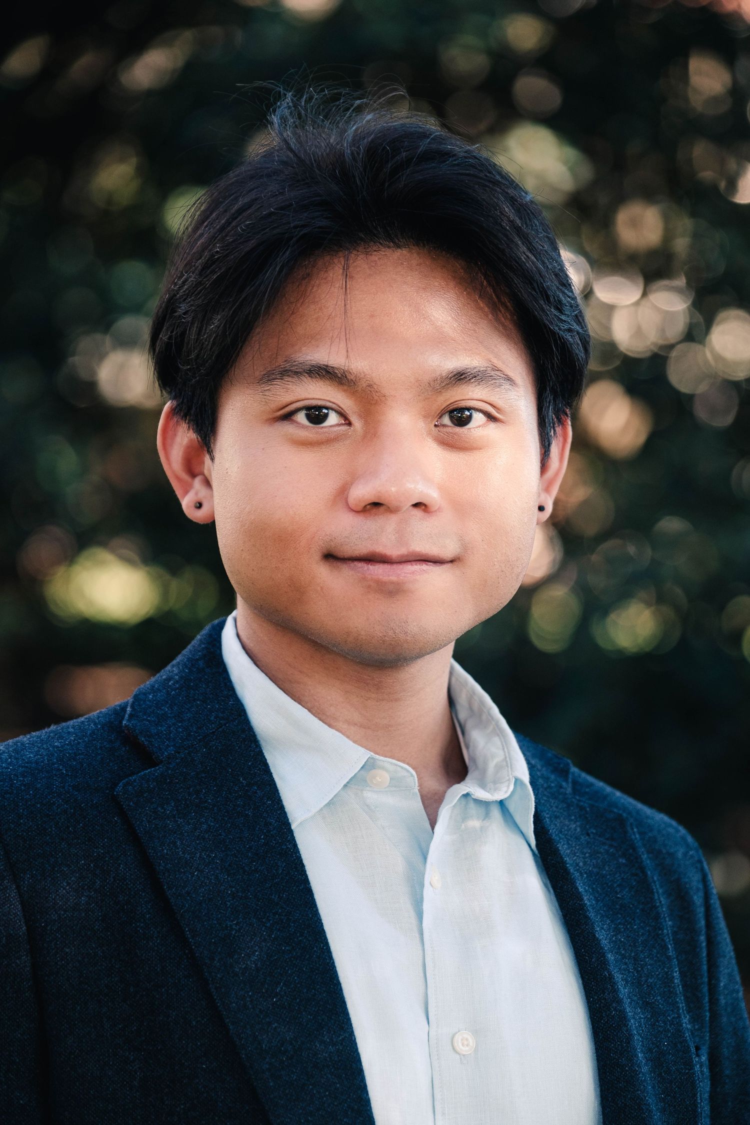 Young man in dark blazer and light shirt smiling outdoors with blurred green trees in background.