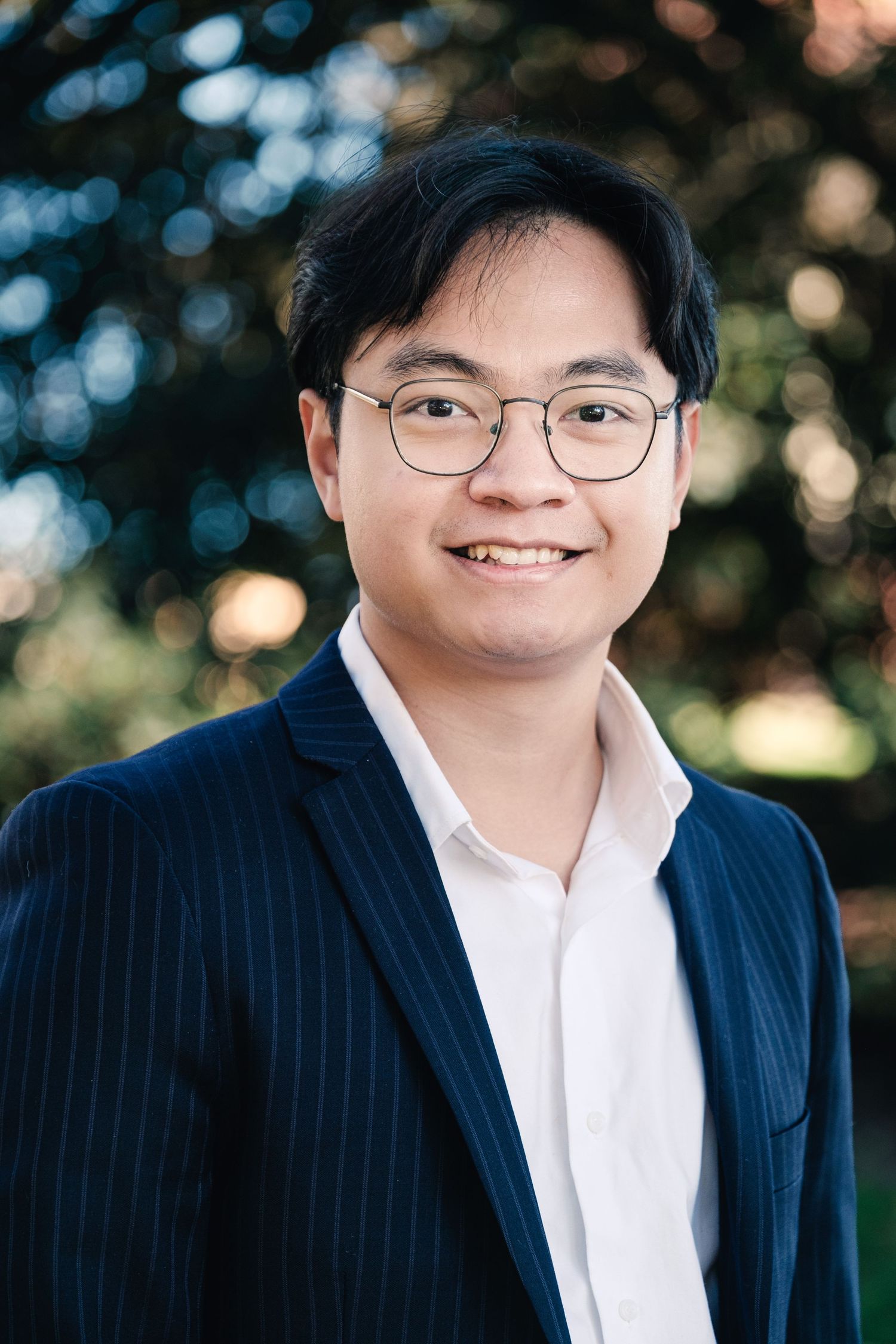 Young Asian man smiling in glasses and navy pinstripe blazer, professional outdoor portrait photo.