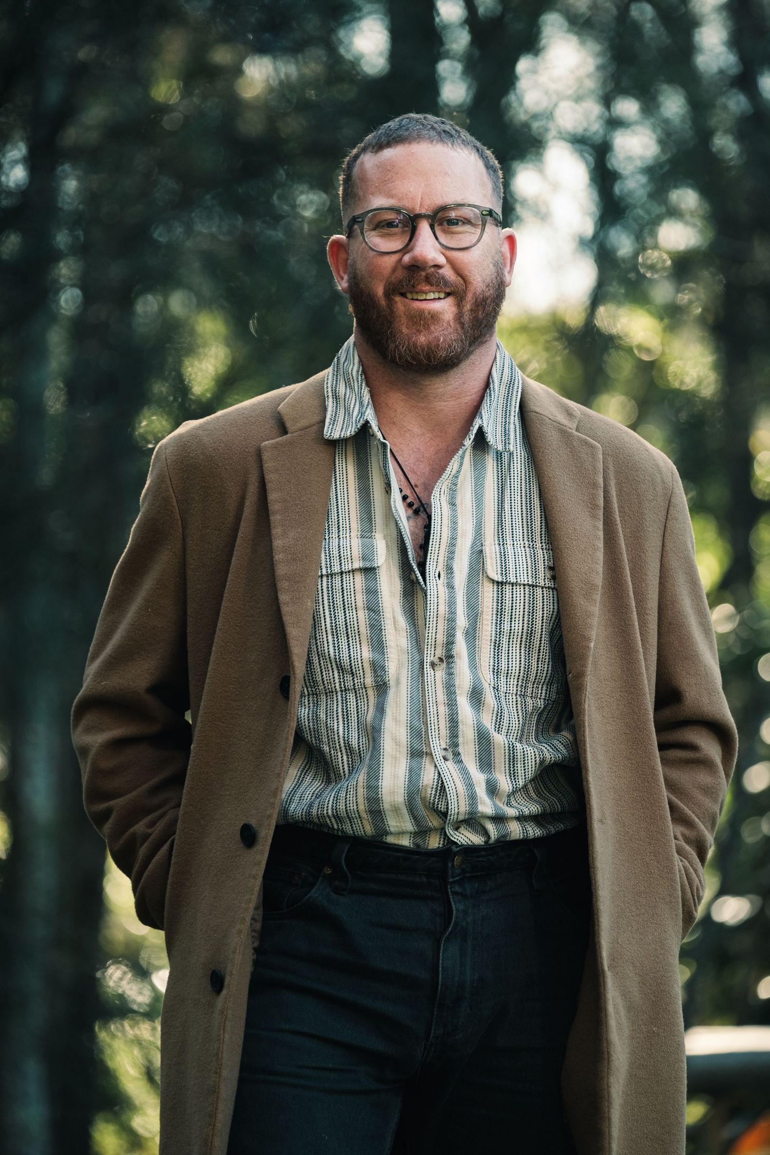 Smiling bearded man wearing glasses, brown overcoat, and striped shirt standing outdoors among green trees.