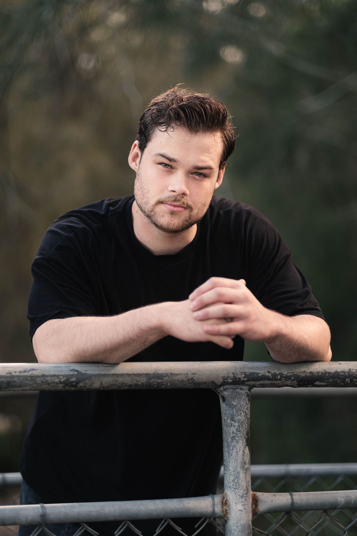 Young man in black t-shirt leaning on metal railing outdoors with blurred green trees in background.