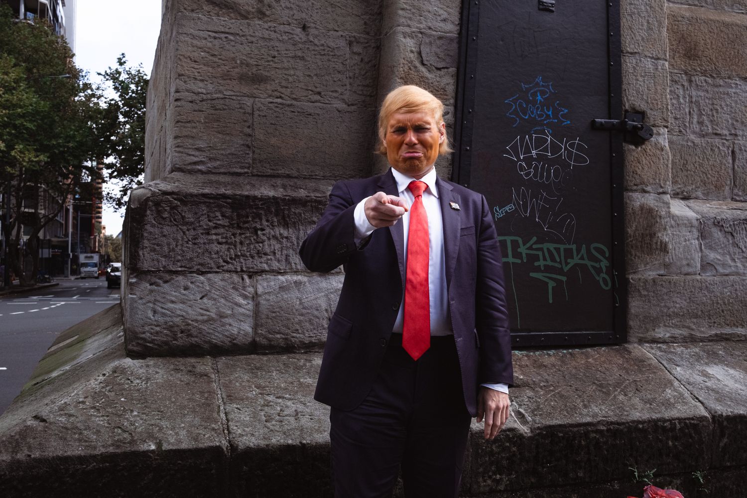 Man in blue suit and red tie points finger while standing near graffiti-covered stone wall outdoors.