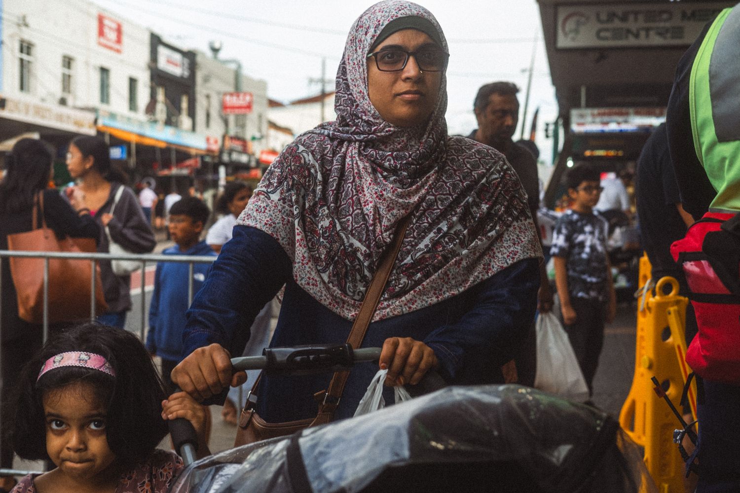 A woman wearing a hijab pushes a stroller through a busy outdoor market with a child nearby.