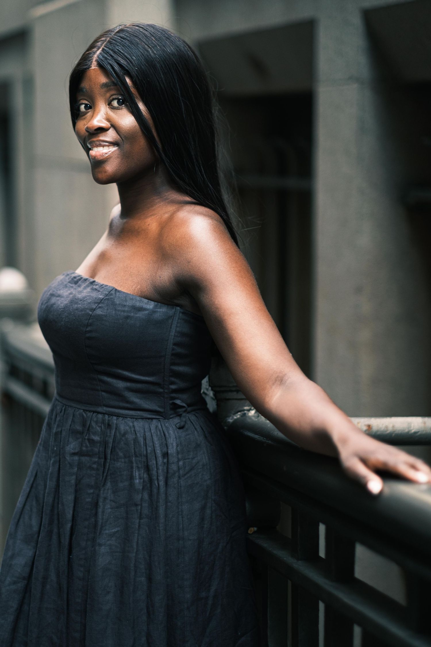 A smiling Black woman in a black strapless dress leans on a railing in an urban setting.