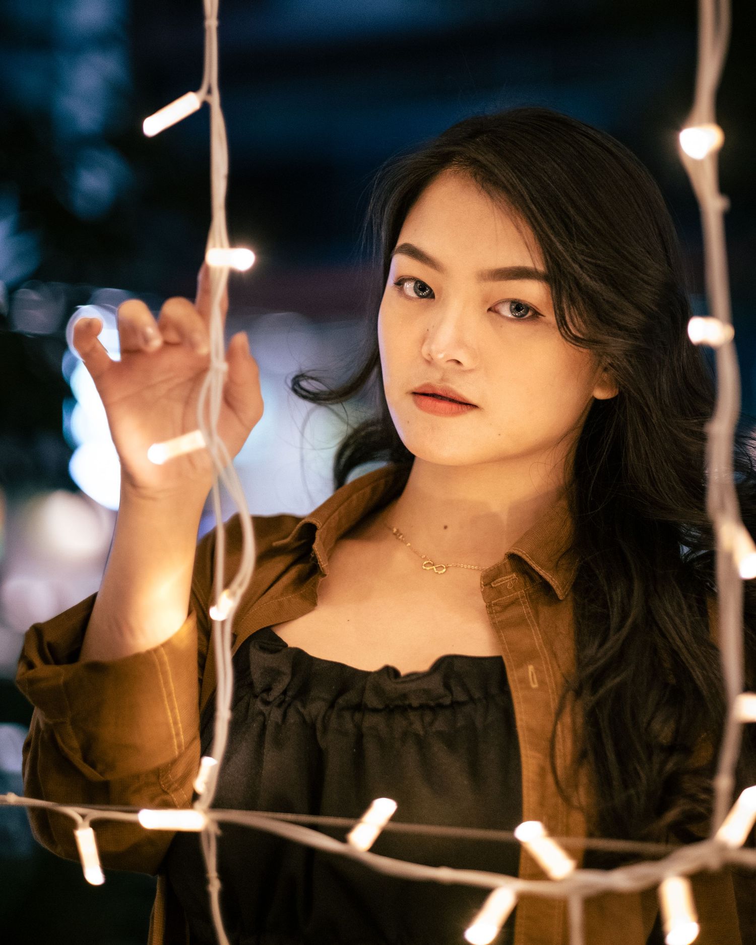A young Asian woman reaches for string lights in a bokeh night photography portrait.