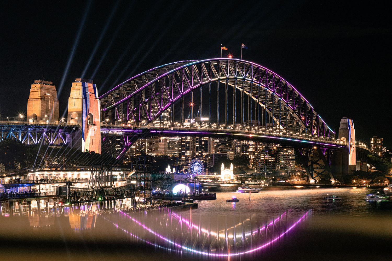 Sydney Harbour Bridge illuminated with colorful lights reflecting on the water at night.