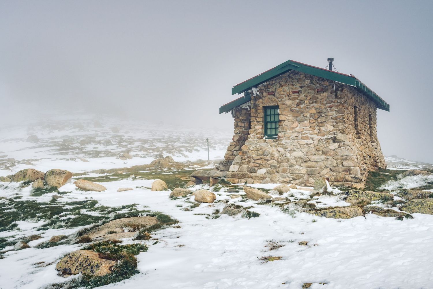 Rustic stone mountain hut covered in snow surrounded by foggy misty alpine winter landscape.