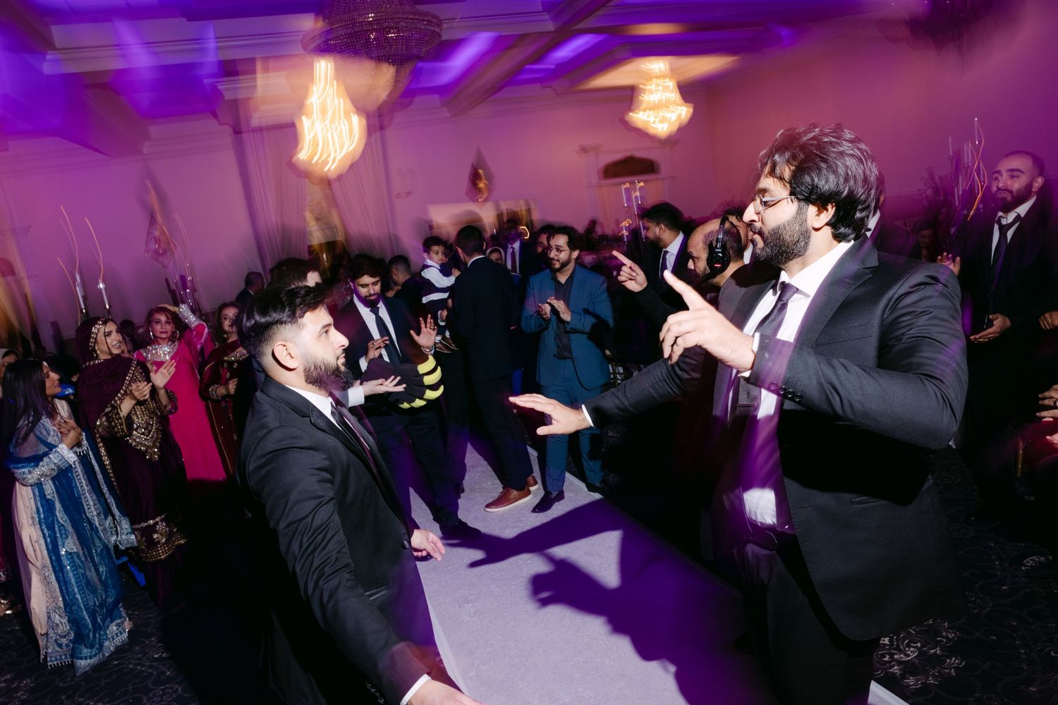 Two men in black suits dancing at a vibrant wedding reception with purple lighting and guests in background.