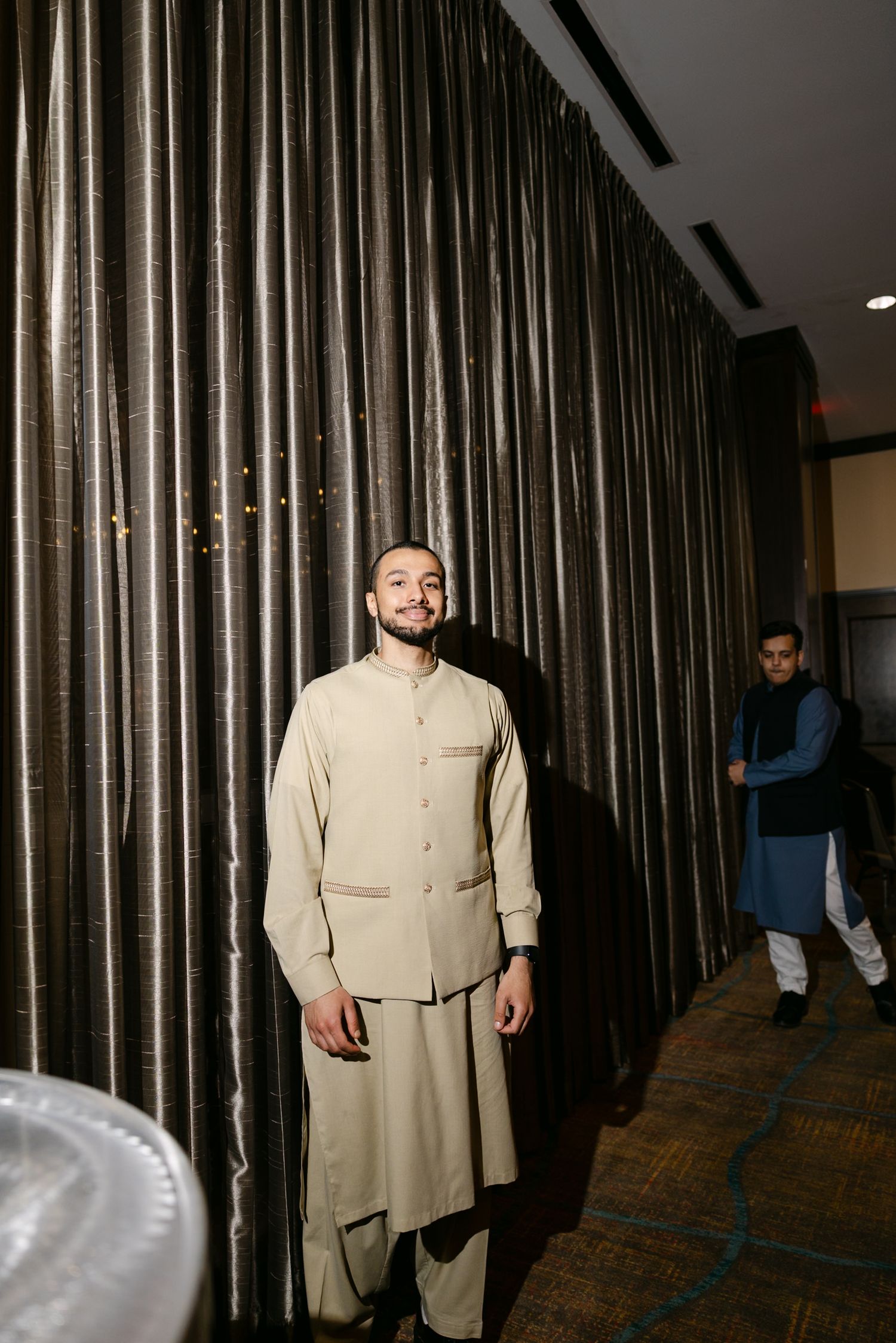 Man in traditional beige South Asian kurta outfit standing near silver curtains at an indoor event.