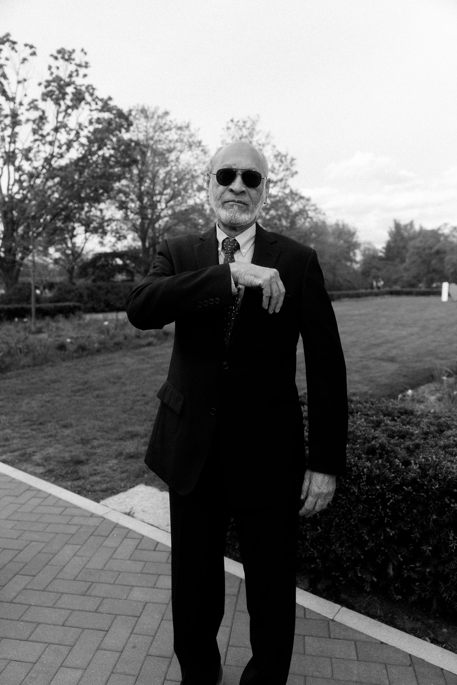 Elderly man in black suit and sunglasses standing on brick path in park, black and white photo.