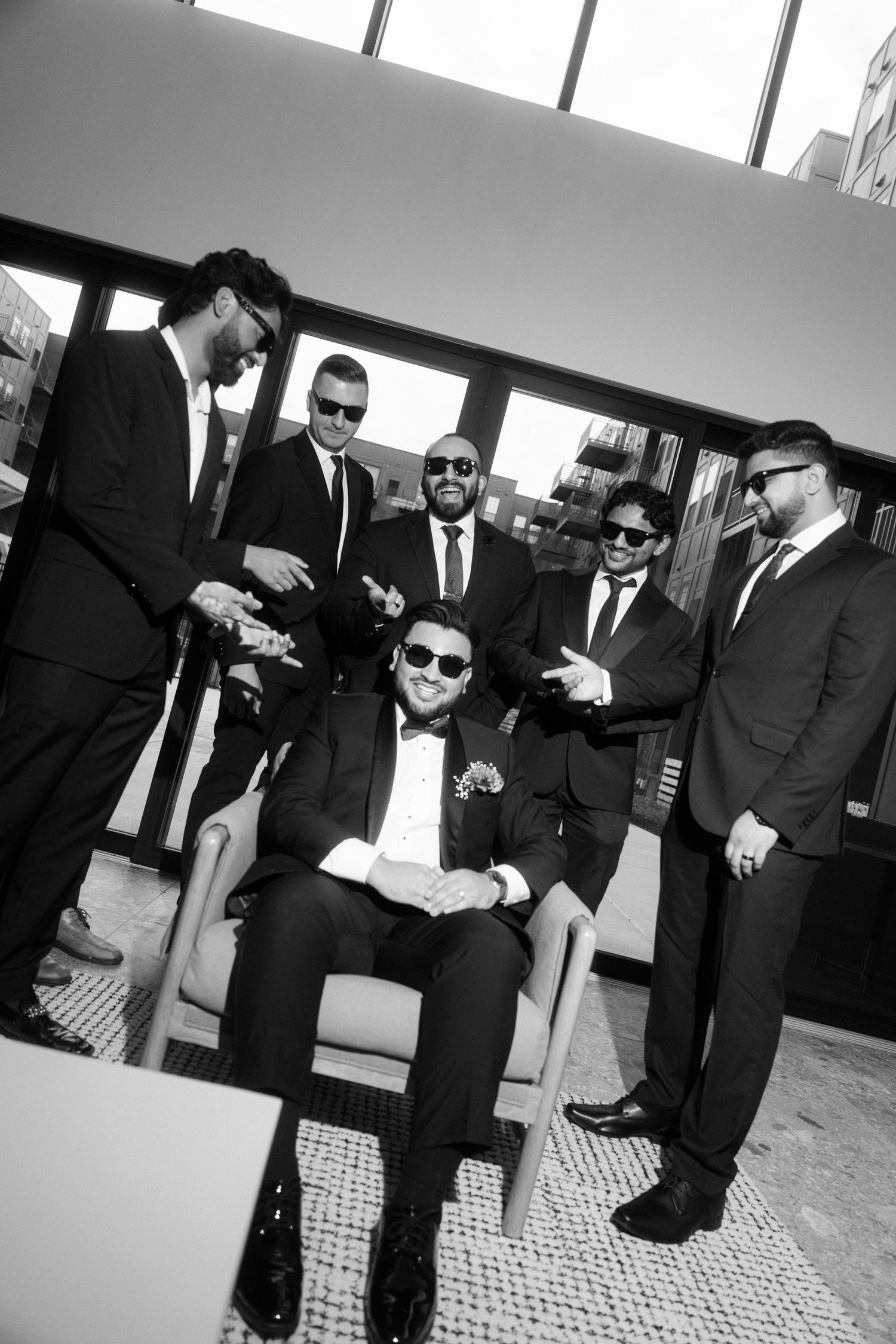 Black and white photo of a groom seated in a chair surrounded by smiling groomsmen in suits on a rooftop.