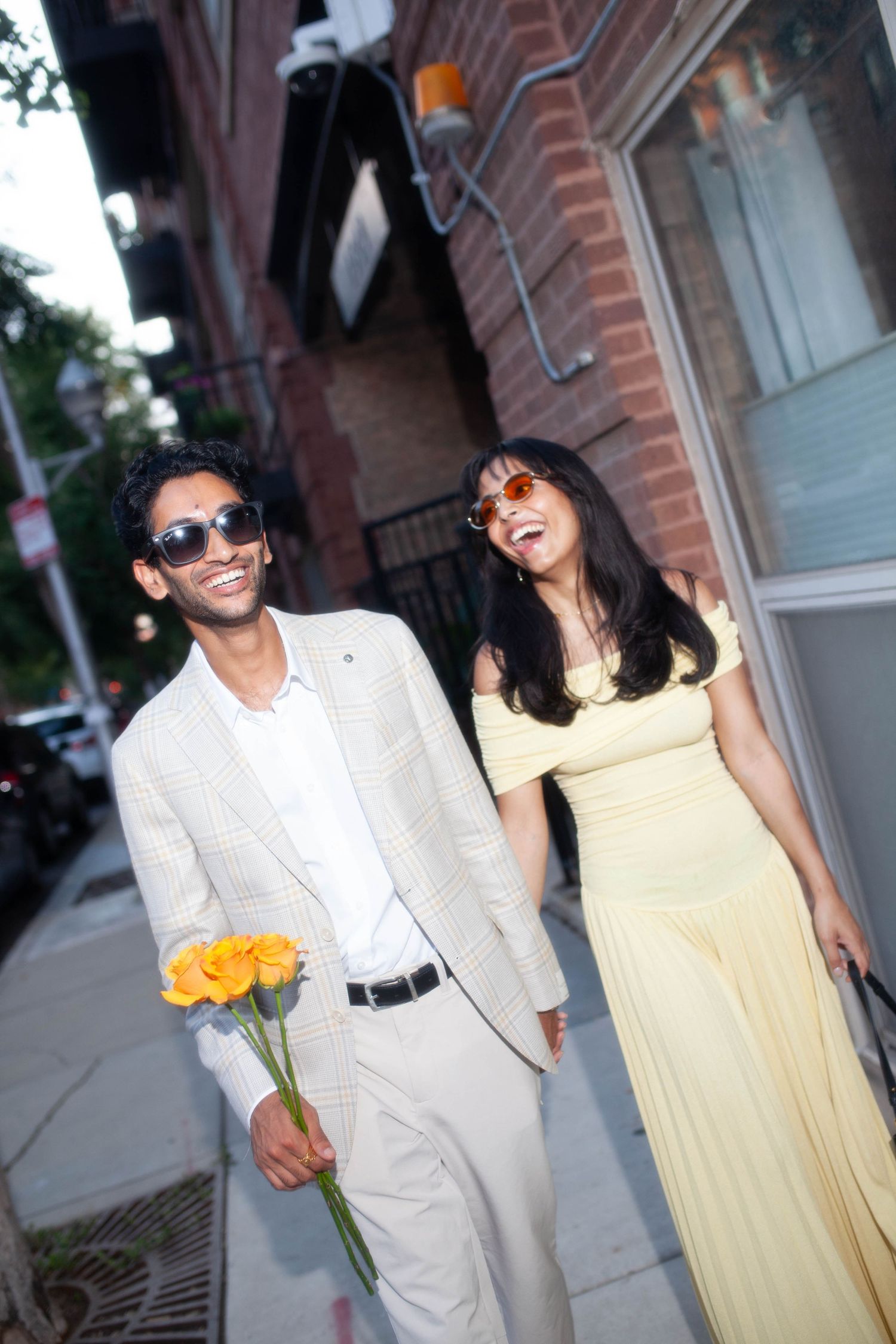 Smiling couple in sunglasses walking on city sidewalk, man in beige suit holding yellow roses, woman in yellow dress.