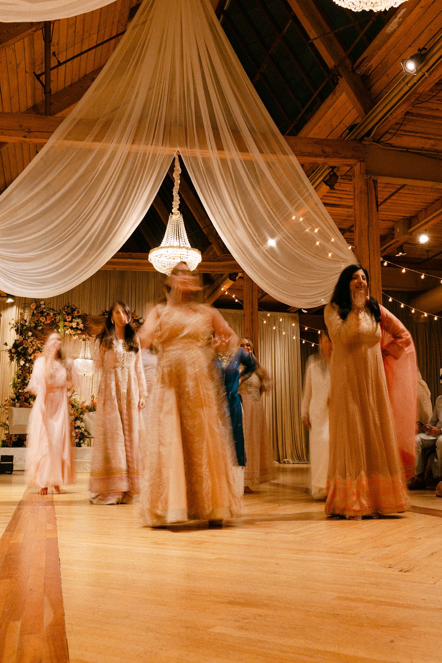 Women in elegant peach and gold lehengas dancing at a decorated wedding reception with draped fabric ceiling.