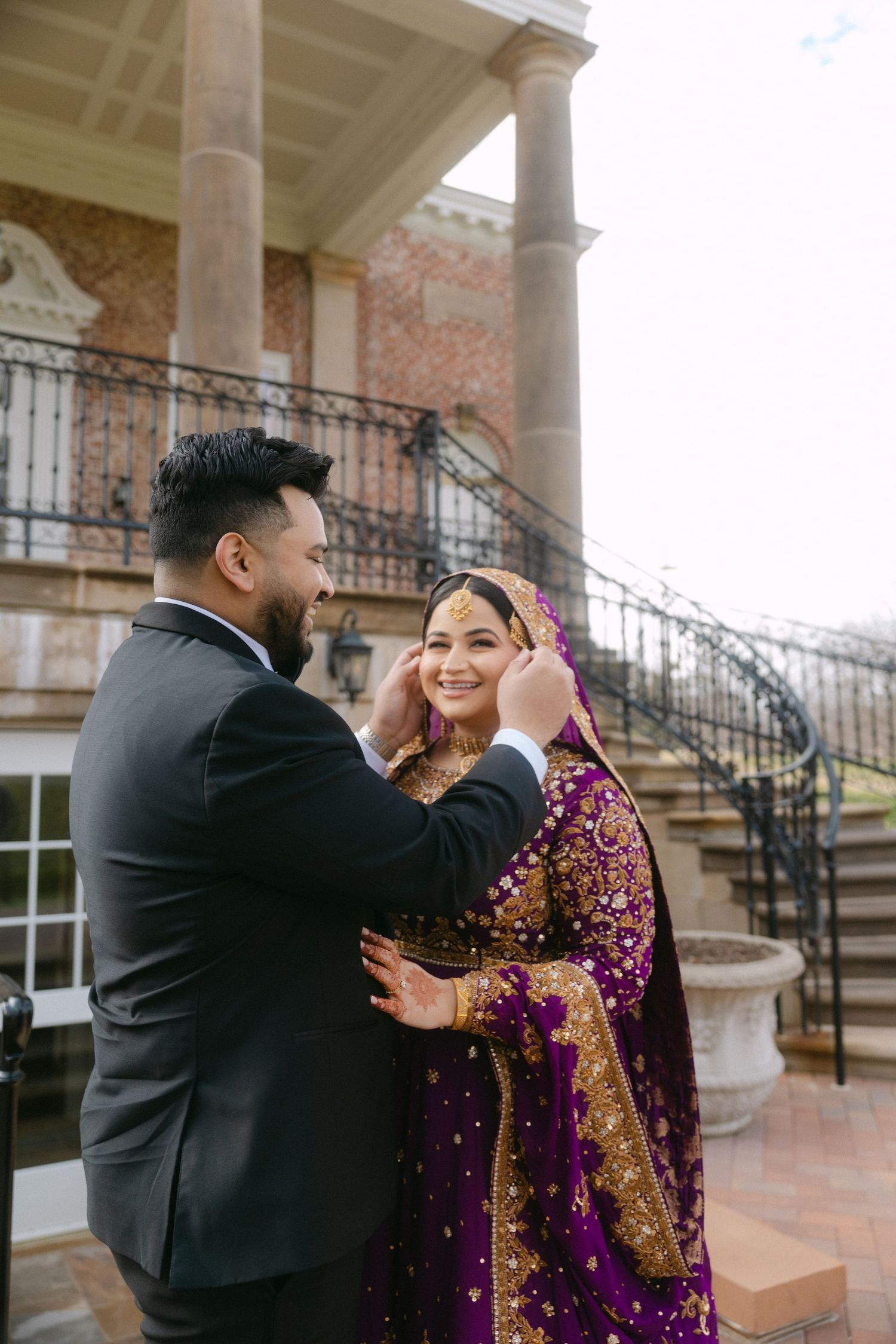 Groom in black suit adjusting bride's purple embroidered dupatta outside elegant venue with grand staircase.