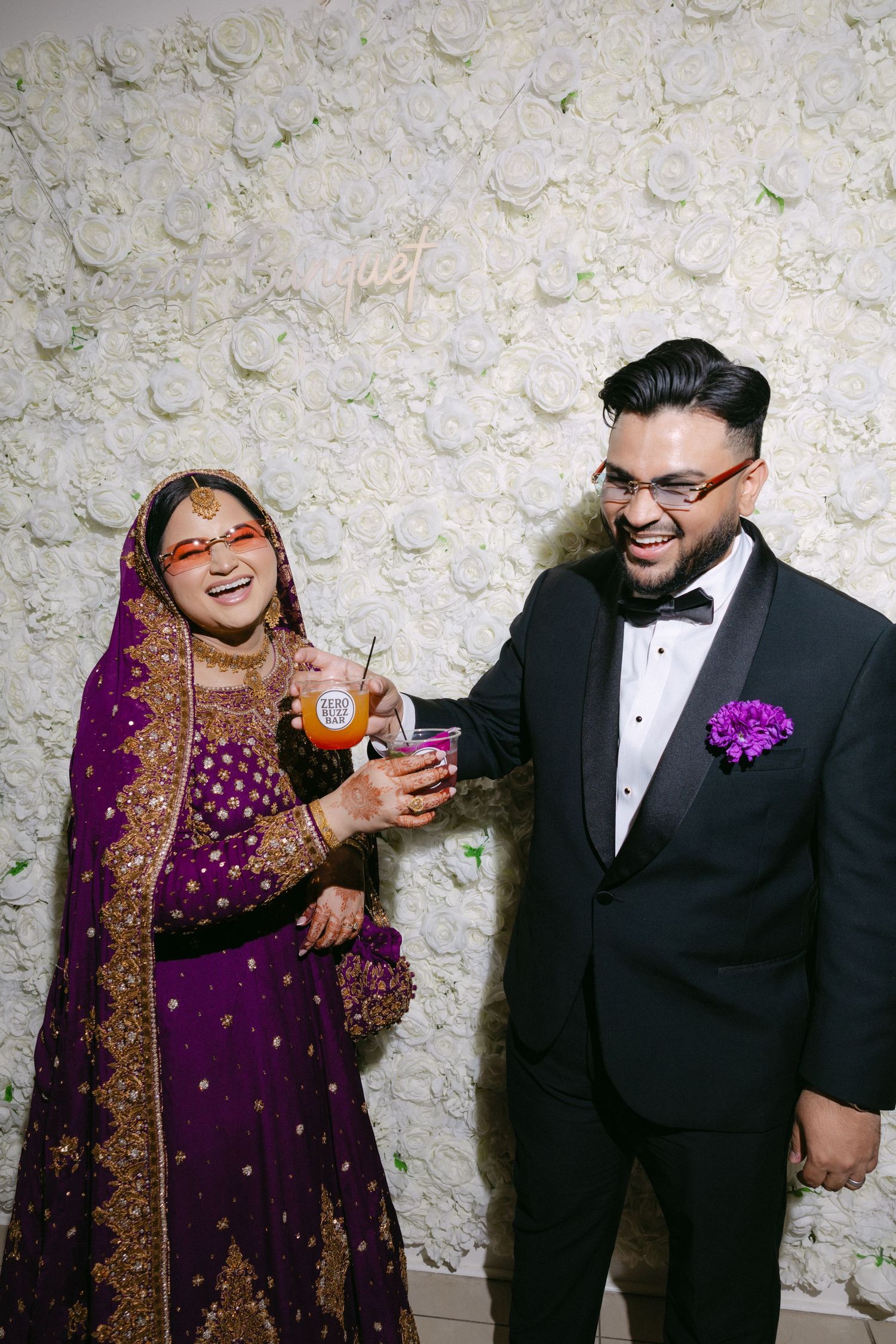 Indian bride in purple lehenga and groom in black tuxedo laughing together at wedding reception.