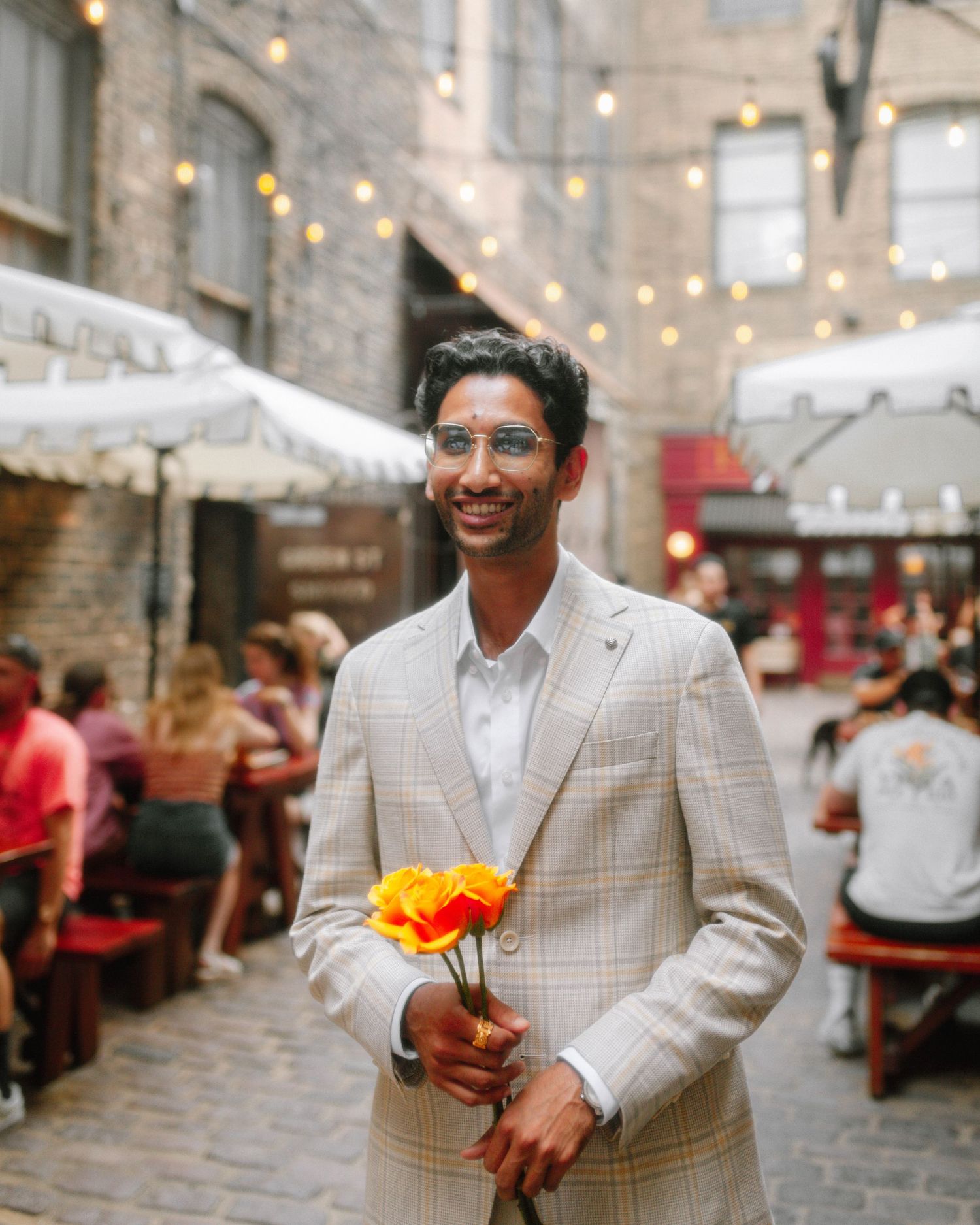 A stylish man in a plaid blazer holds orange flowers in a charming outdoor alley with string lights.