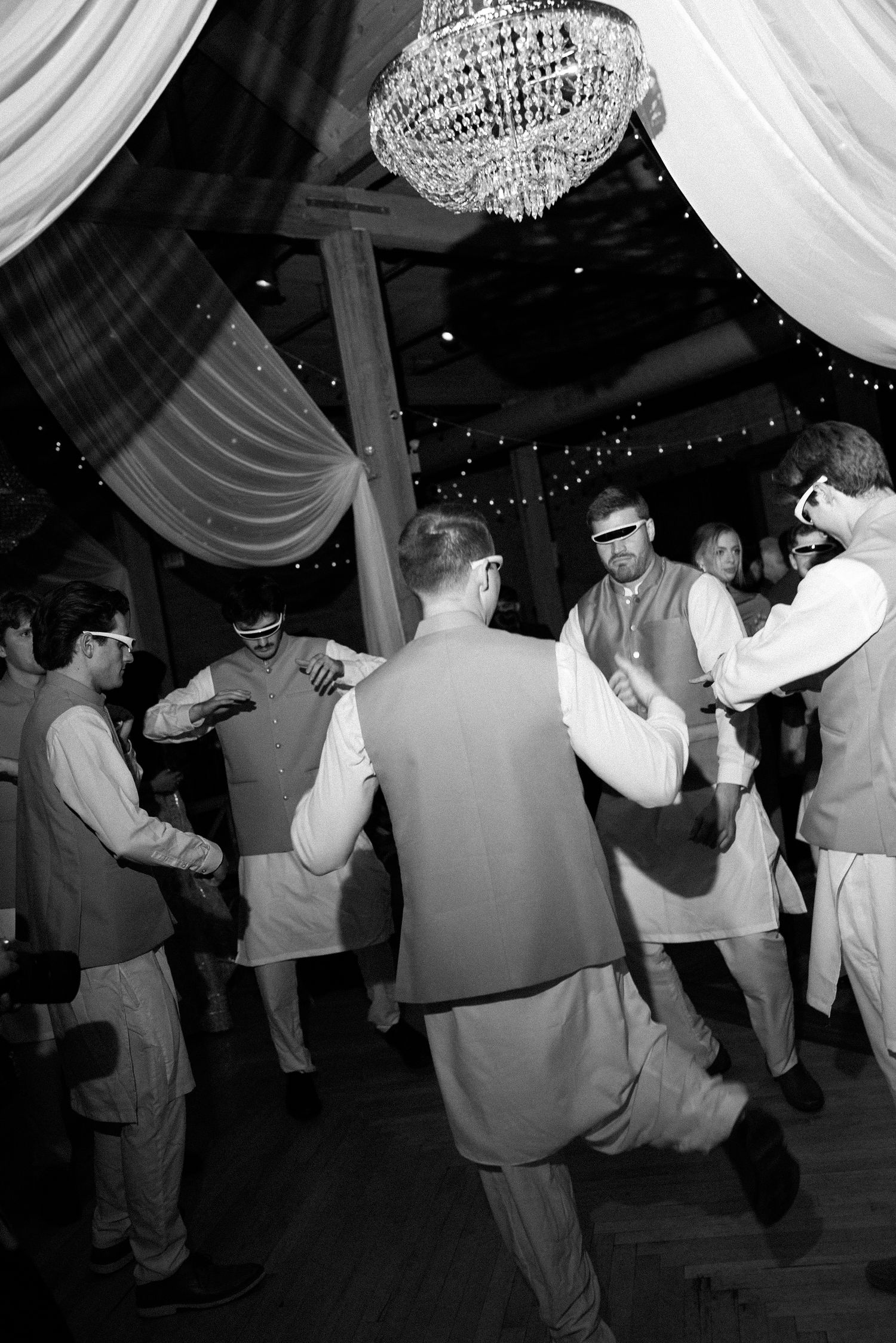 Black and white photo of men in traditional South Asian attire dancing at a wedding reception under a chandelier.
