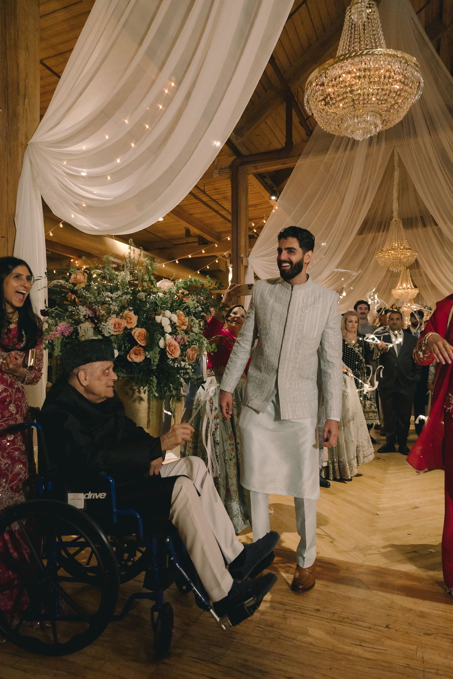 Groom in white sherwani walks beside elderly man in wheelchair at elegant Indian wedding reception with chandelier.