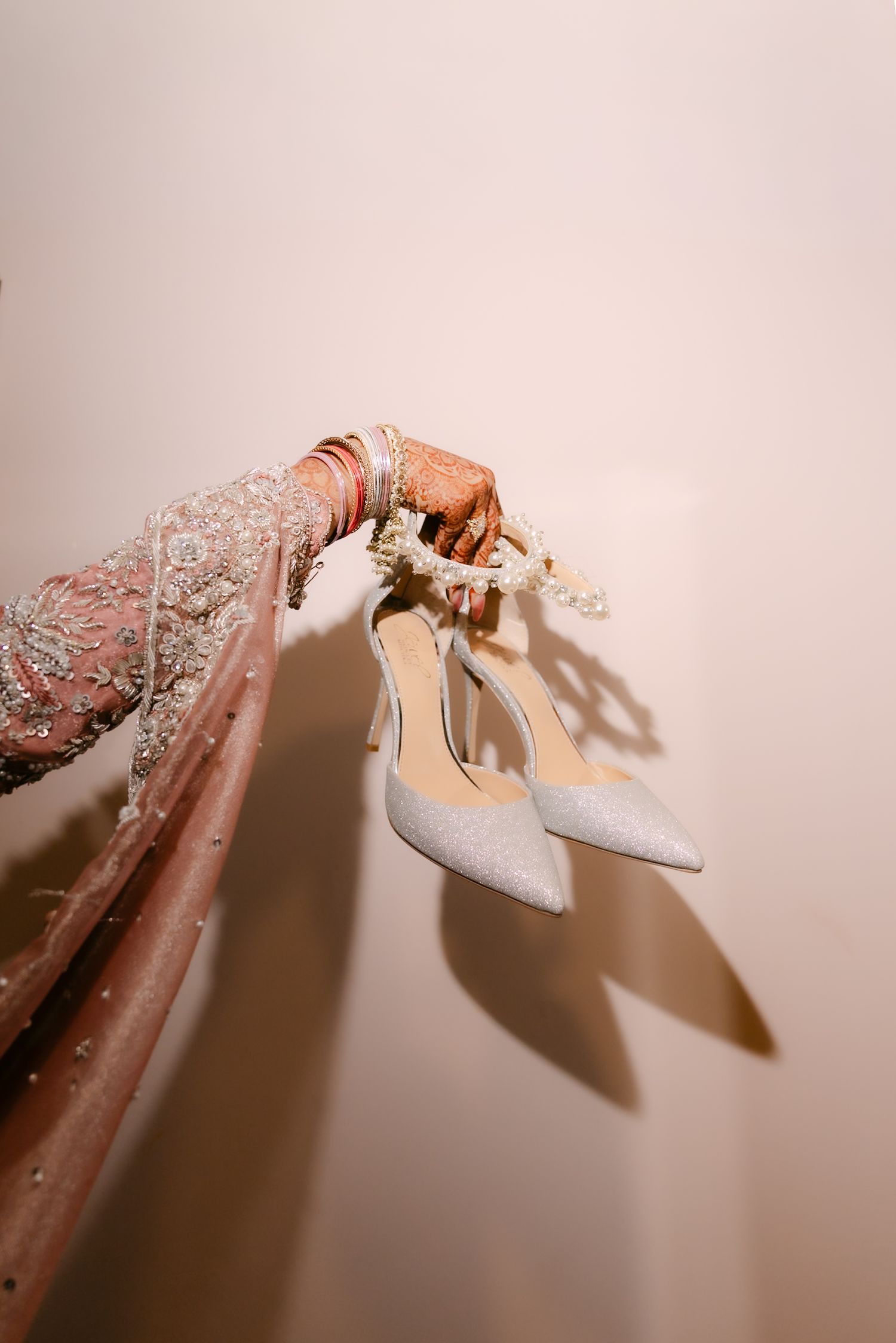 Bridal hand with mehndi and bangles holding silver glitter heels against a neutral background.