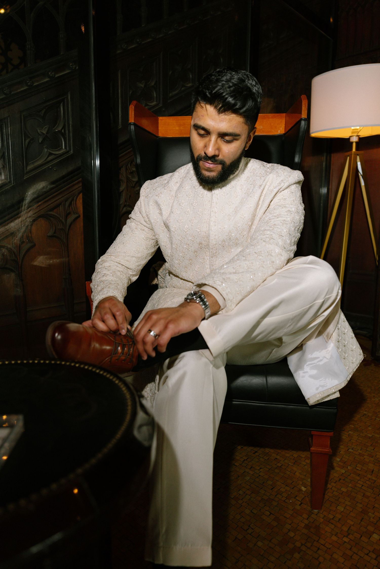 Man in white embroidered sherwani sitting on chair adjusting brown leather shoes in elegant dark interior.