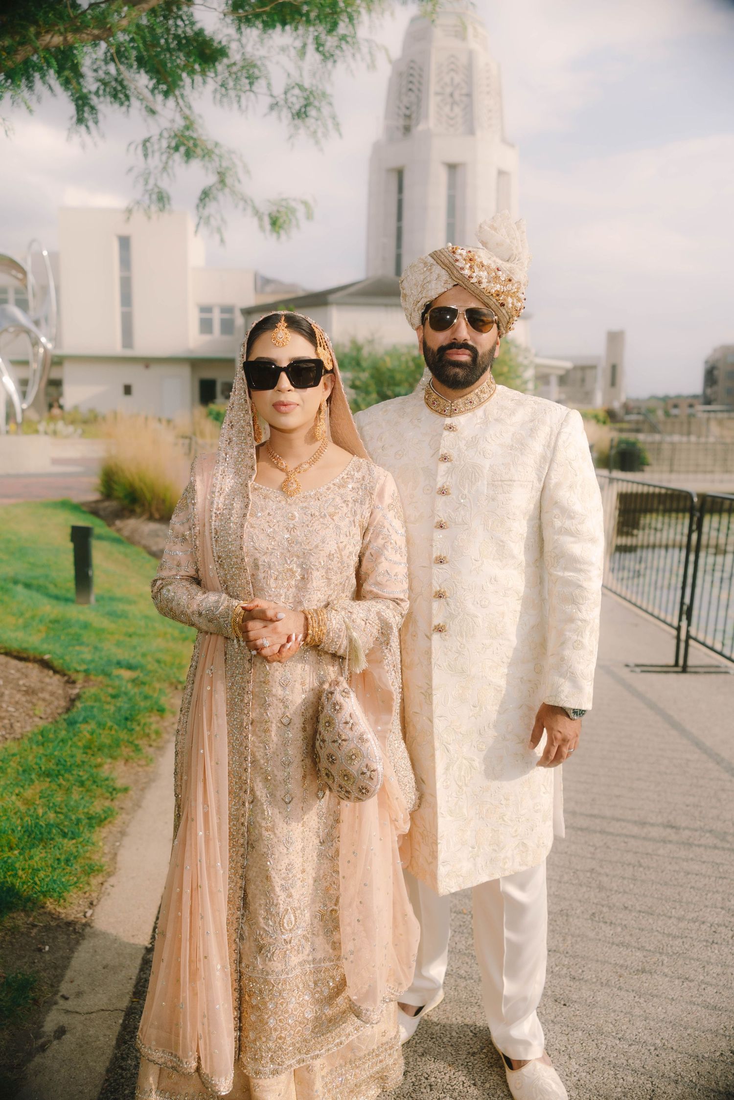 South Asian couple in matching ivory wedding attire with sunglasses pose outdoors near a white building.