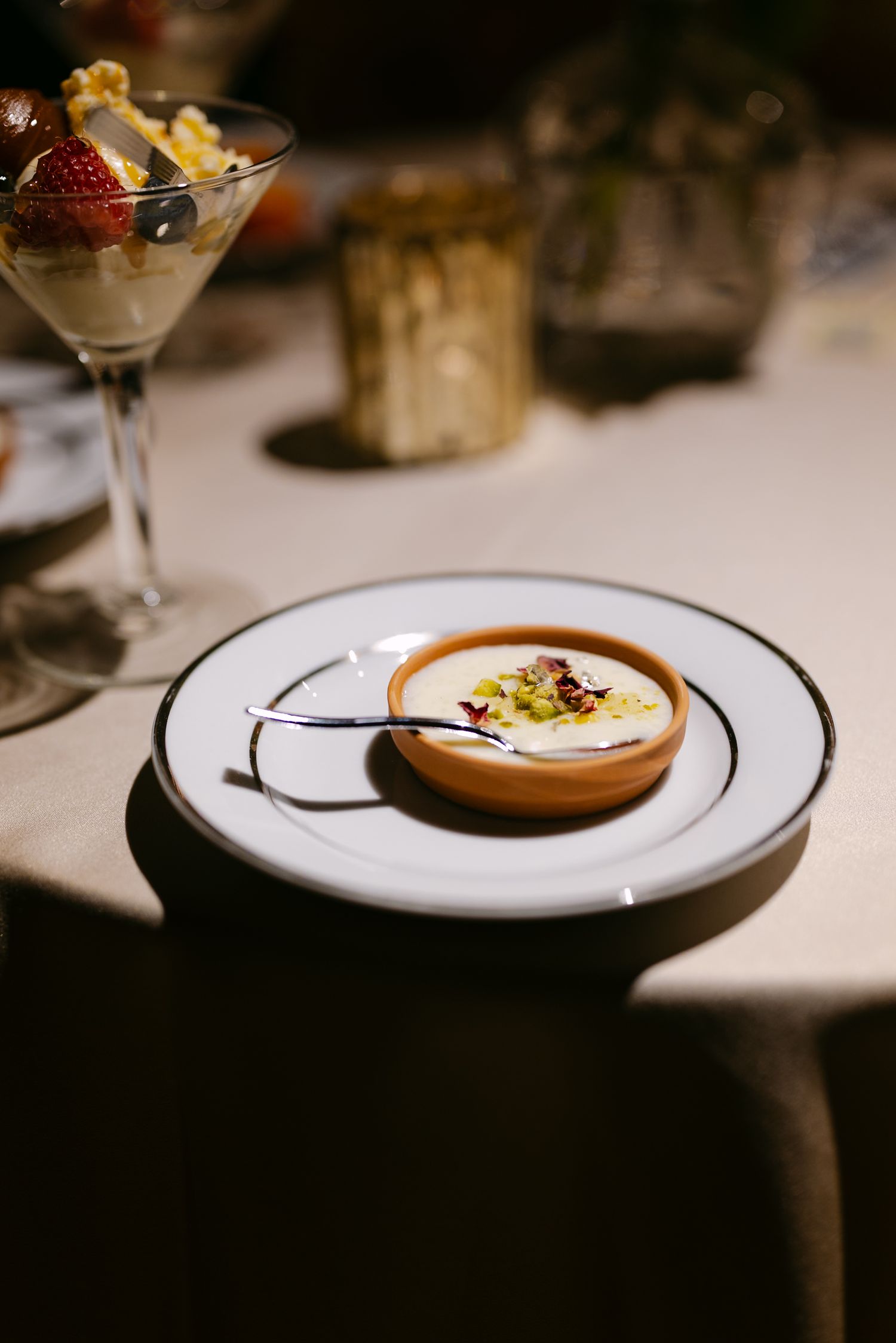 Elegant plated dessert in a terracotta bowl on white china, garnished with pistachios, beside a martini glass with strawberries.