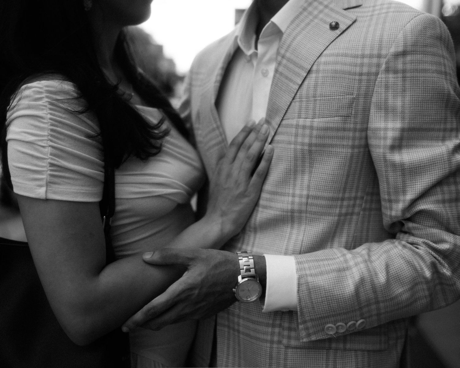Black and white photo of a couple embracing, woman in white dress touching man in plaid suit wearing a watch.