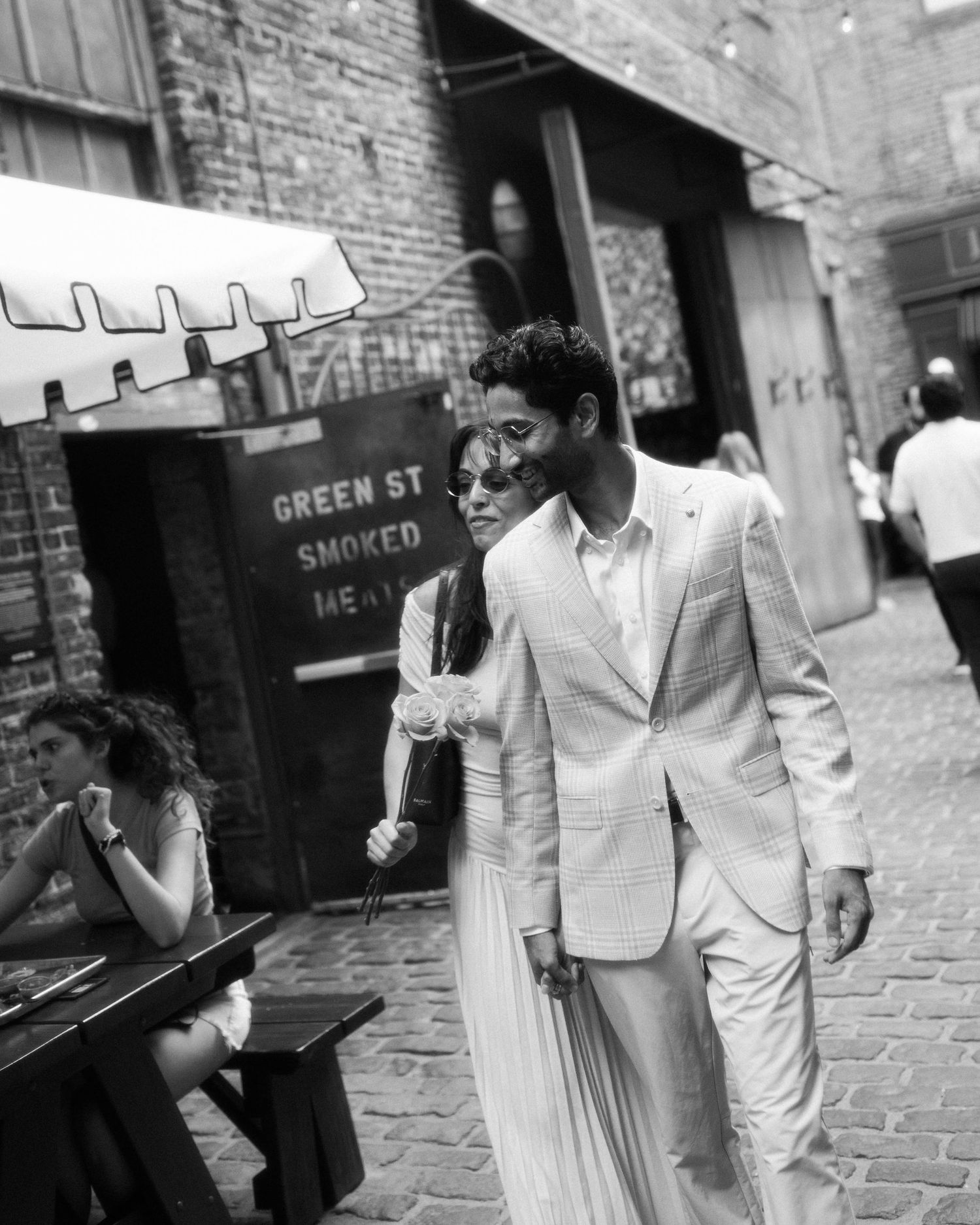 Black and white photo of a couple in formal attire walking past Green St Smoked Meat restaurant in an urban alley.