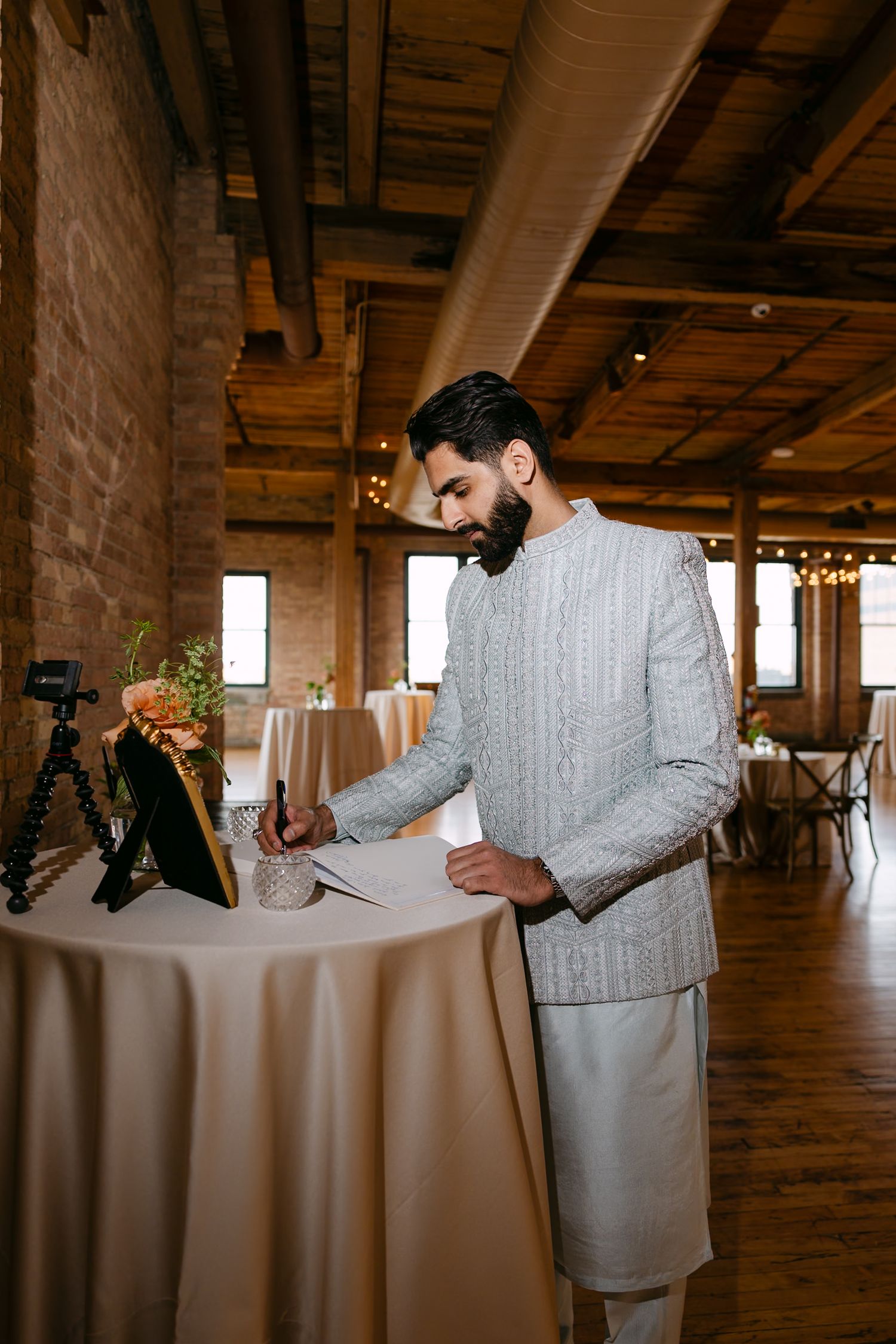 A bearded man in traditional Indian attire signs documents at a decorated table in an elegant rustic wedding venue.