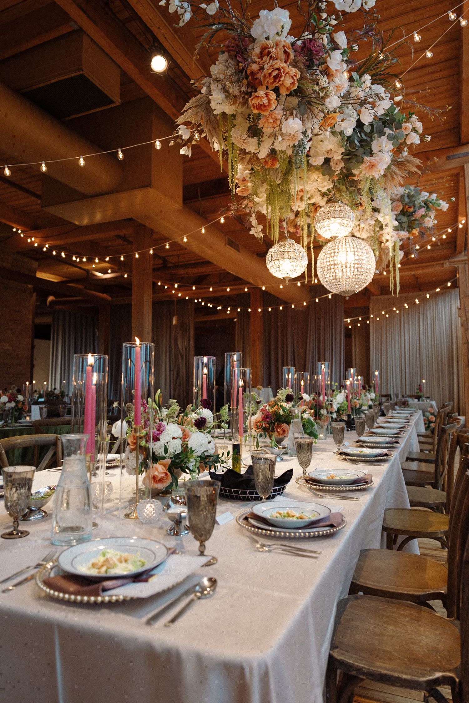 Elegant wedding reception table with floral chandelier, string lights, candles, and formal place settings in a rustic venue.