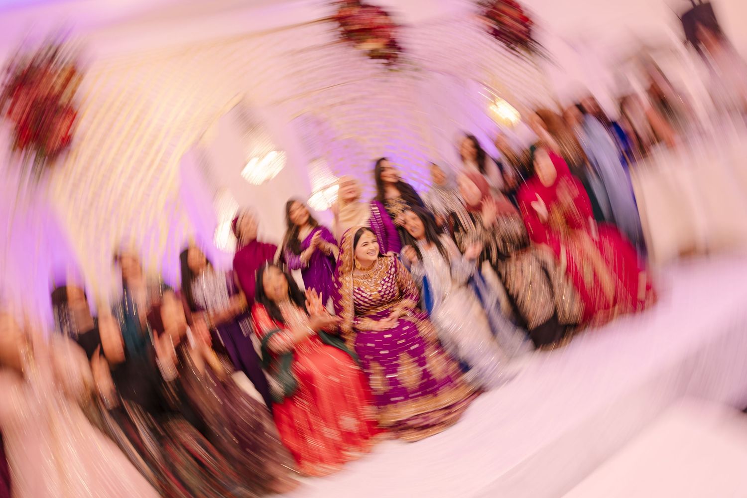 Bride in purple lehenga dancing at Indian Pakistani wedding reception with guests celebrating in blurred motion background.