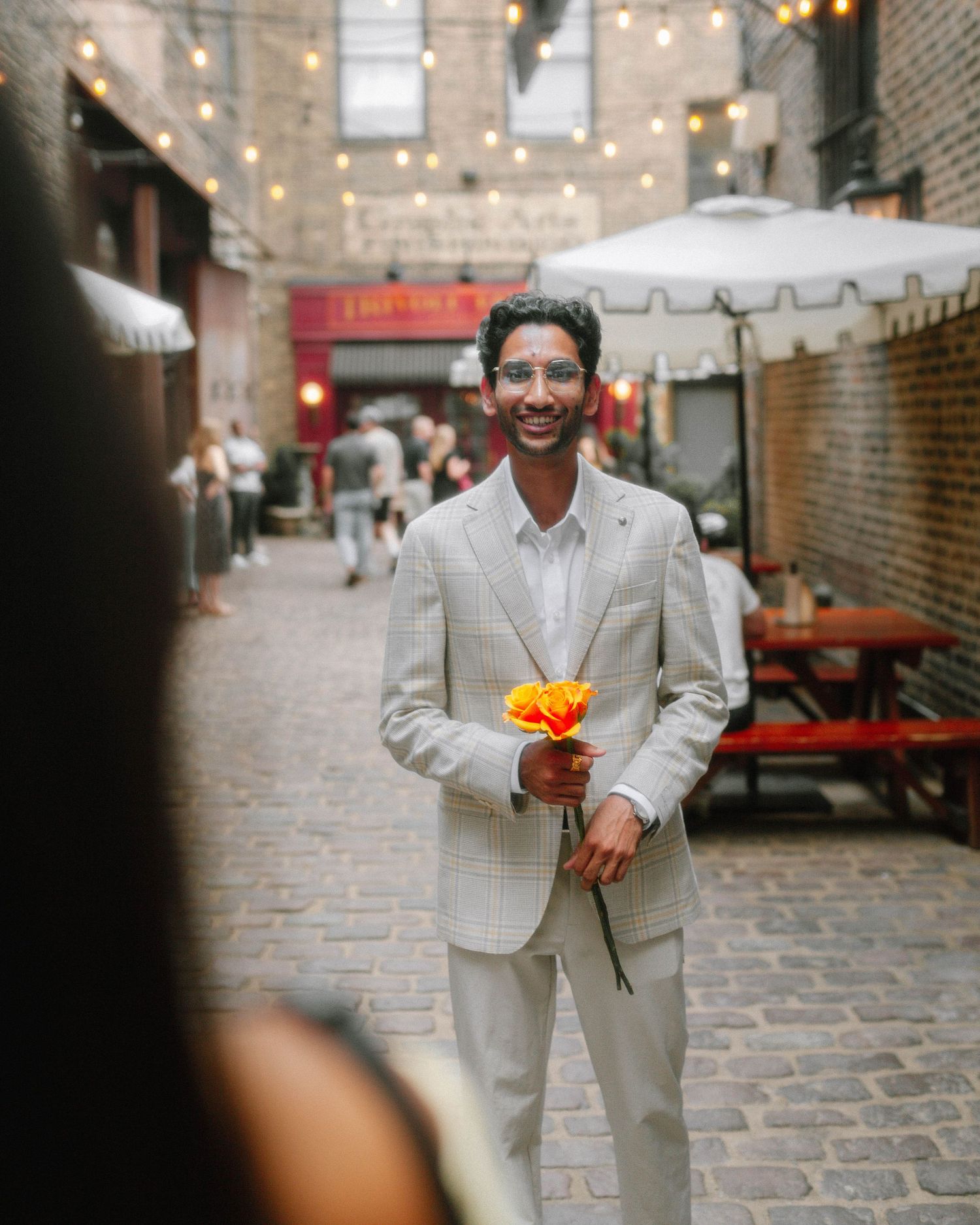 A smiling man in a light gray suit holds orange and yellow flowers on a cobblestone alley with string lights overhead.