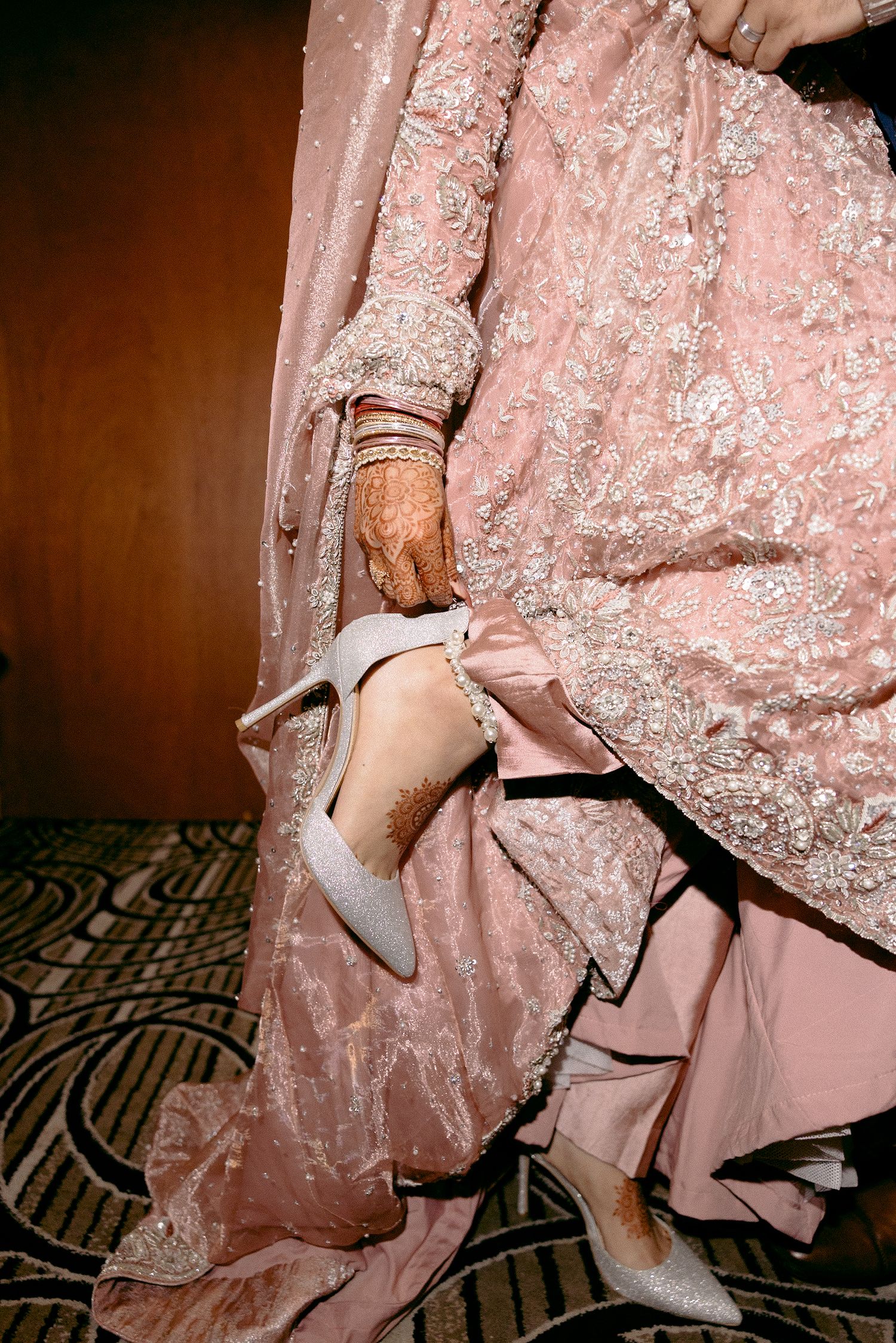 Bride in pink embroidered lehenga with mehndi hands putting on silver heels for South Asian wedding.