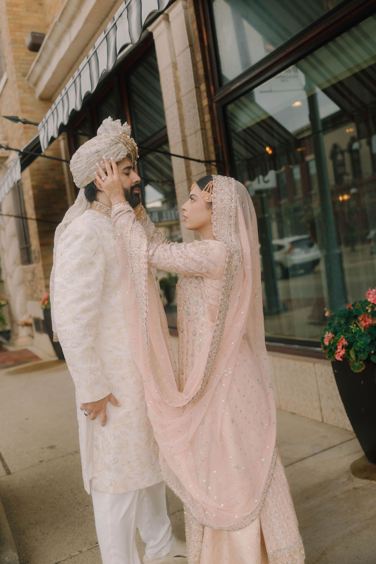South Asian couple in traditional white and blush wedding attire share a tender moment on a city sidewalk.