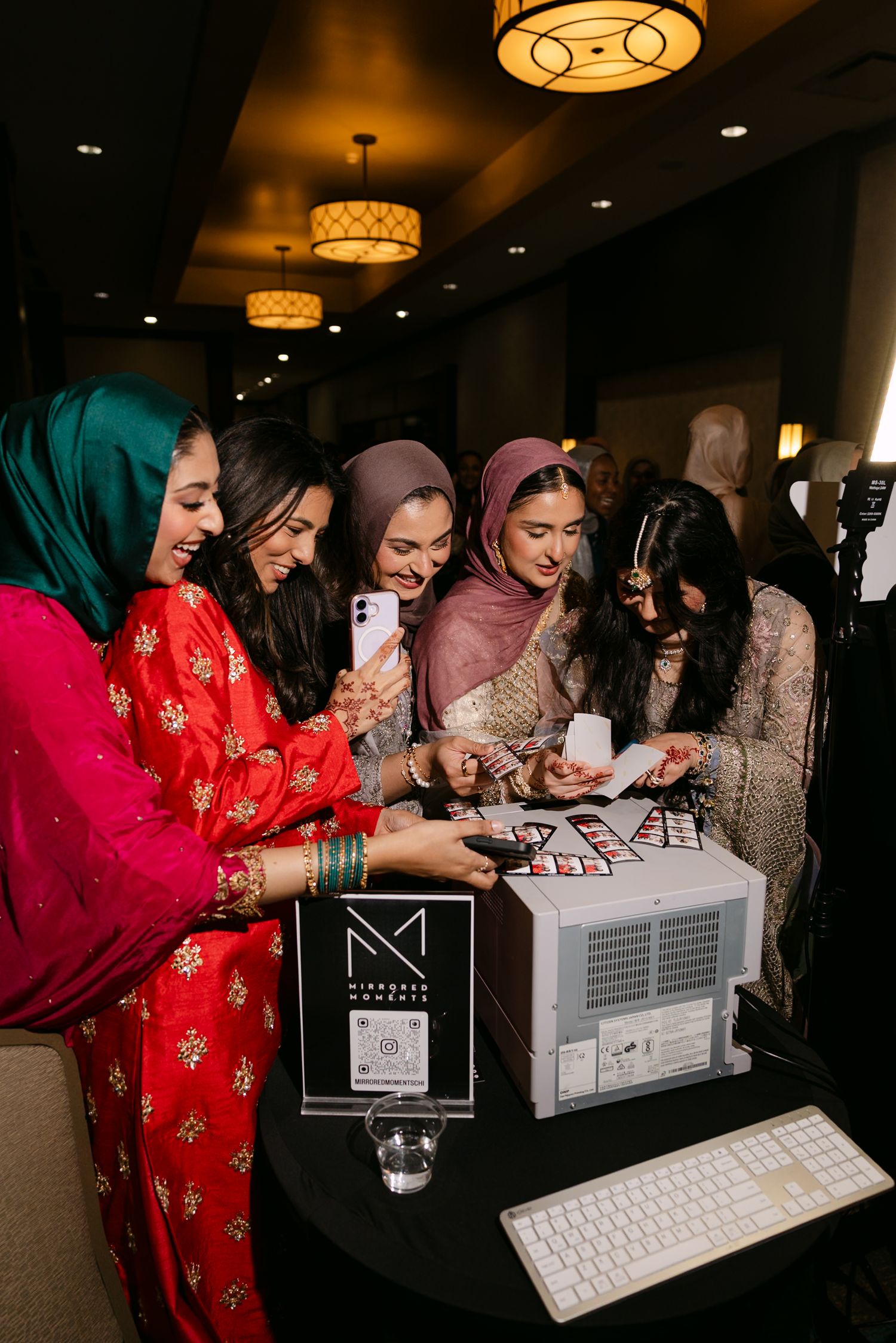 Group of women in hijabs gathered around a photo booth printer at an elegant indoor event venue.