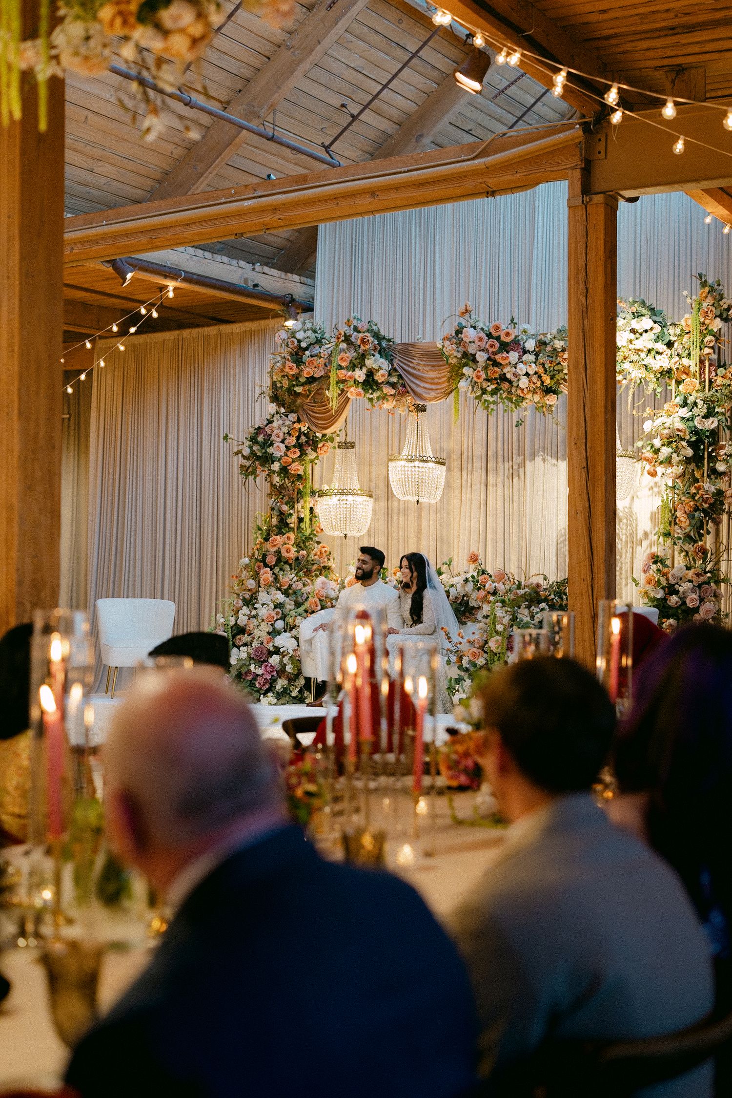 Elegant wedding reception with floral arch backdrop, candlelit tables, and couple seated at sweetheart table in decorated venue.