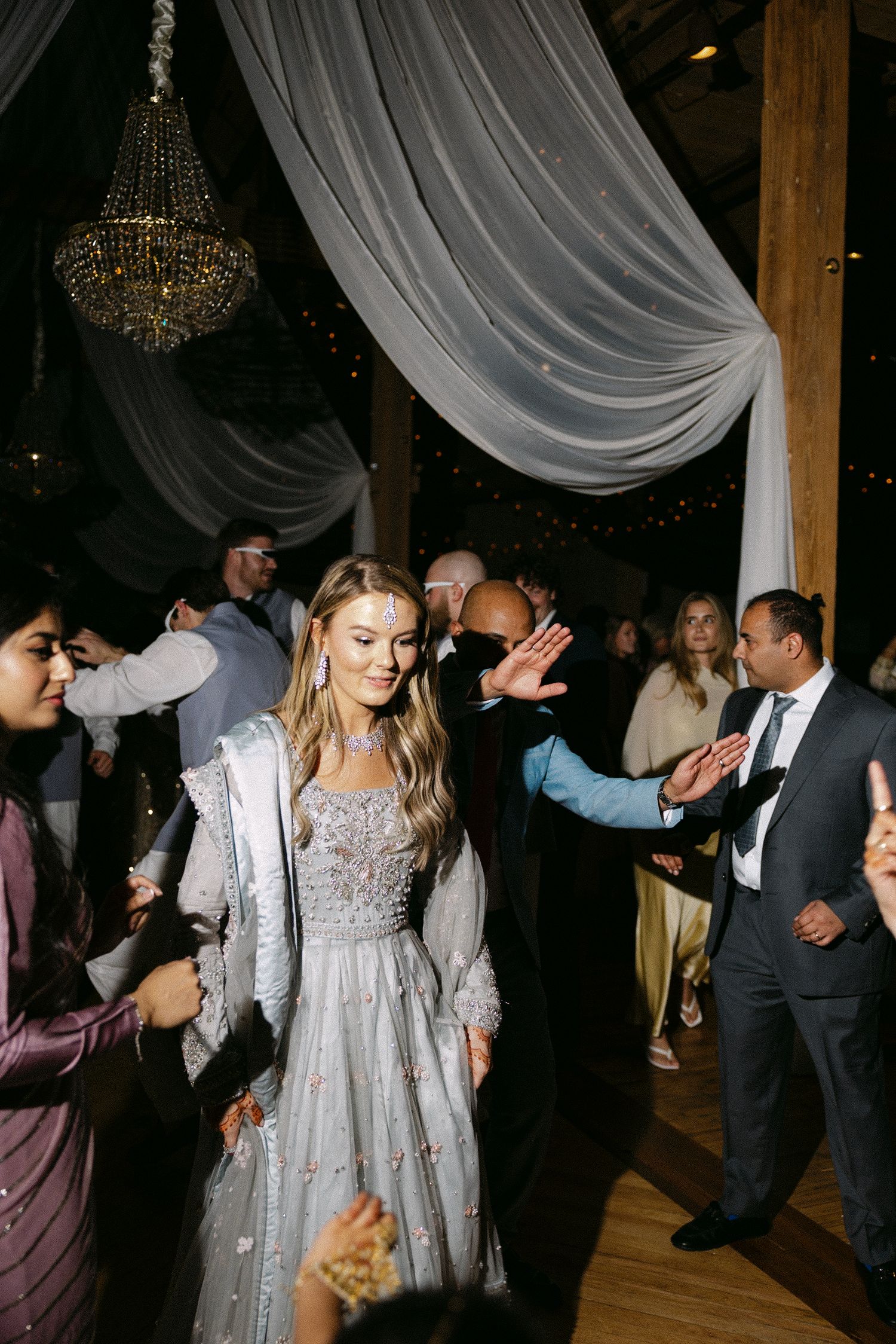 Bride in silver gown walks through guest tunnel at elegant barn wedding reception with chandelier.