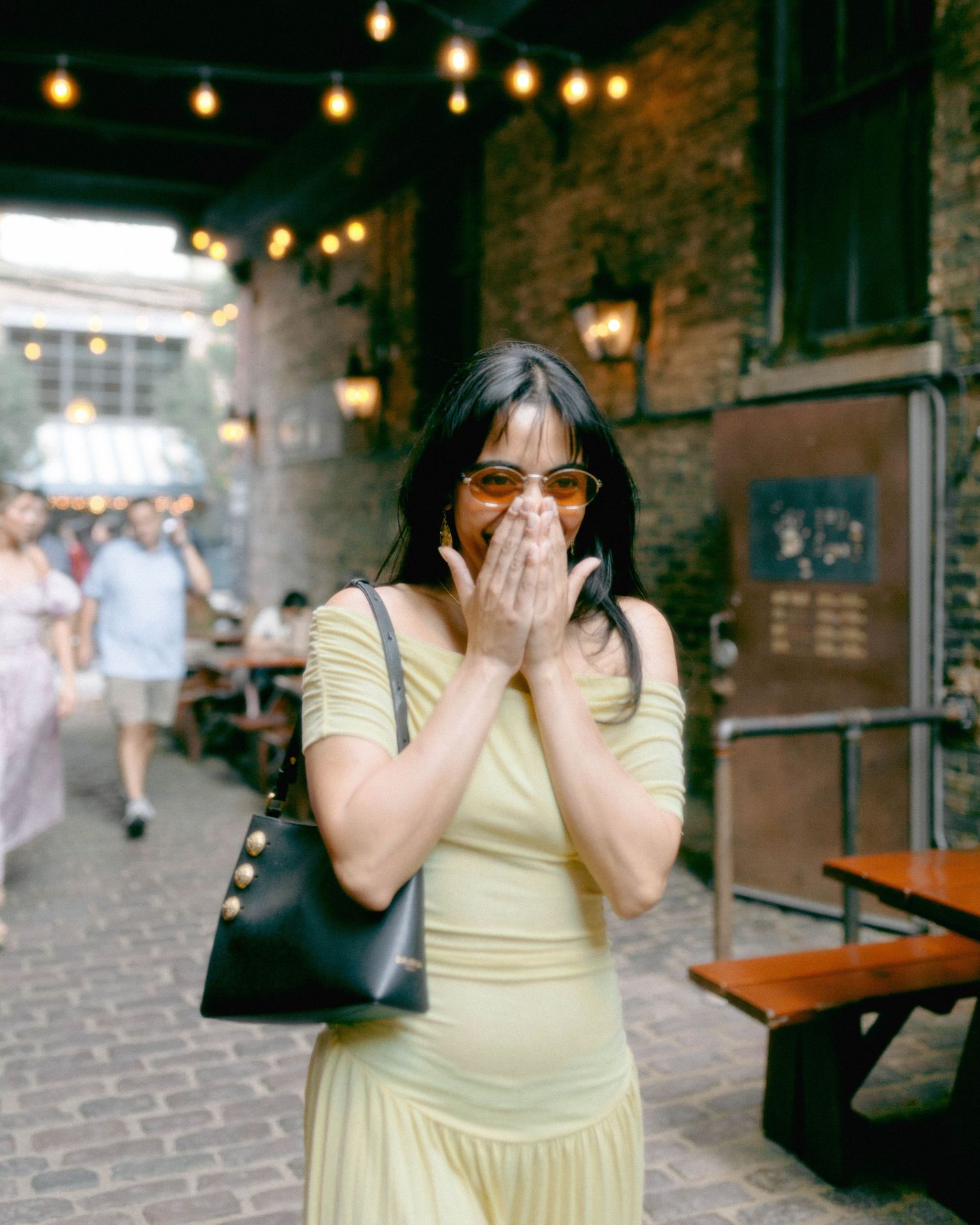Woman in yellow dress with sunglasses covering mouth in surprise, standing in a brick courtyard with string lights.