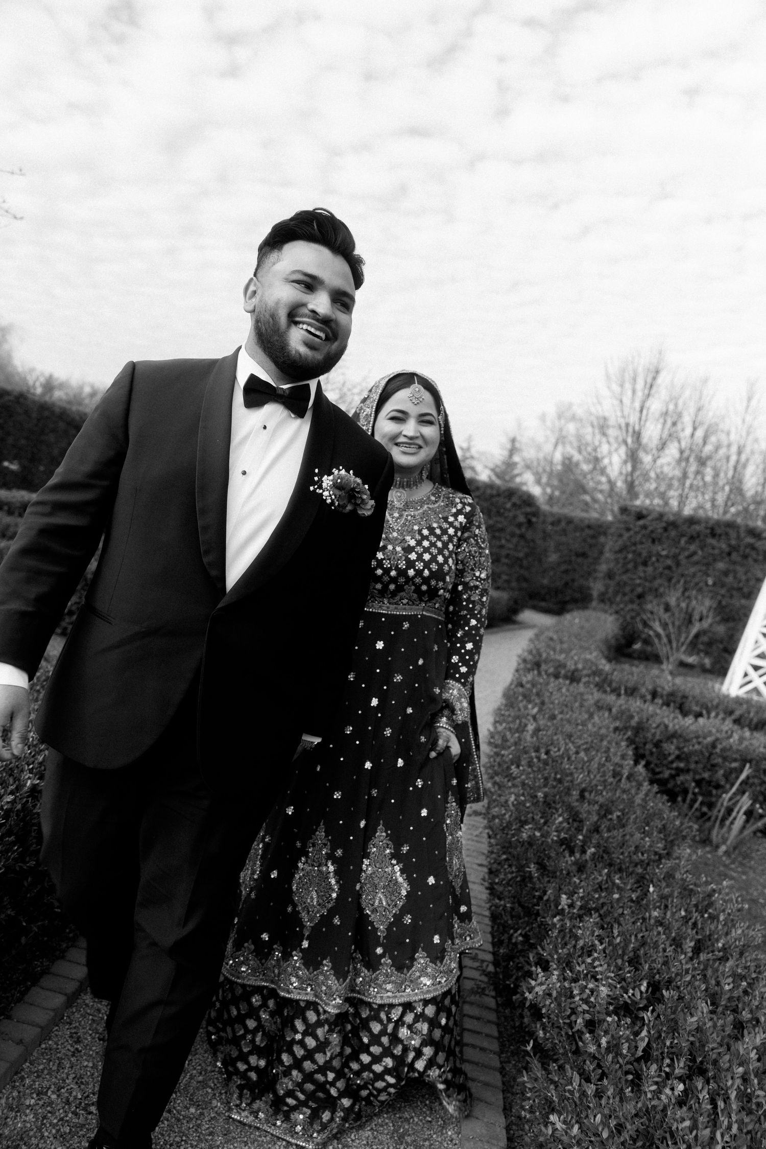 Black and white photo of a smiling couple in formal attire posing in a garden, man in tuxedo, woman in embroidered lehenga.