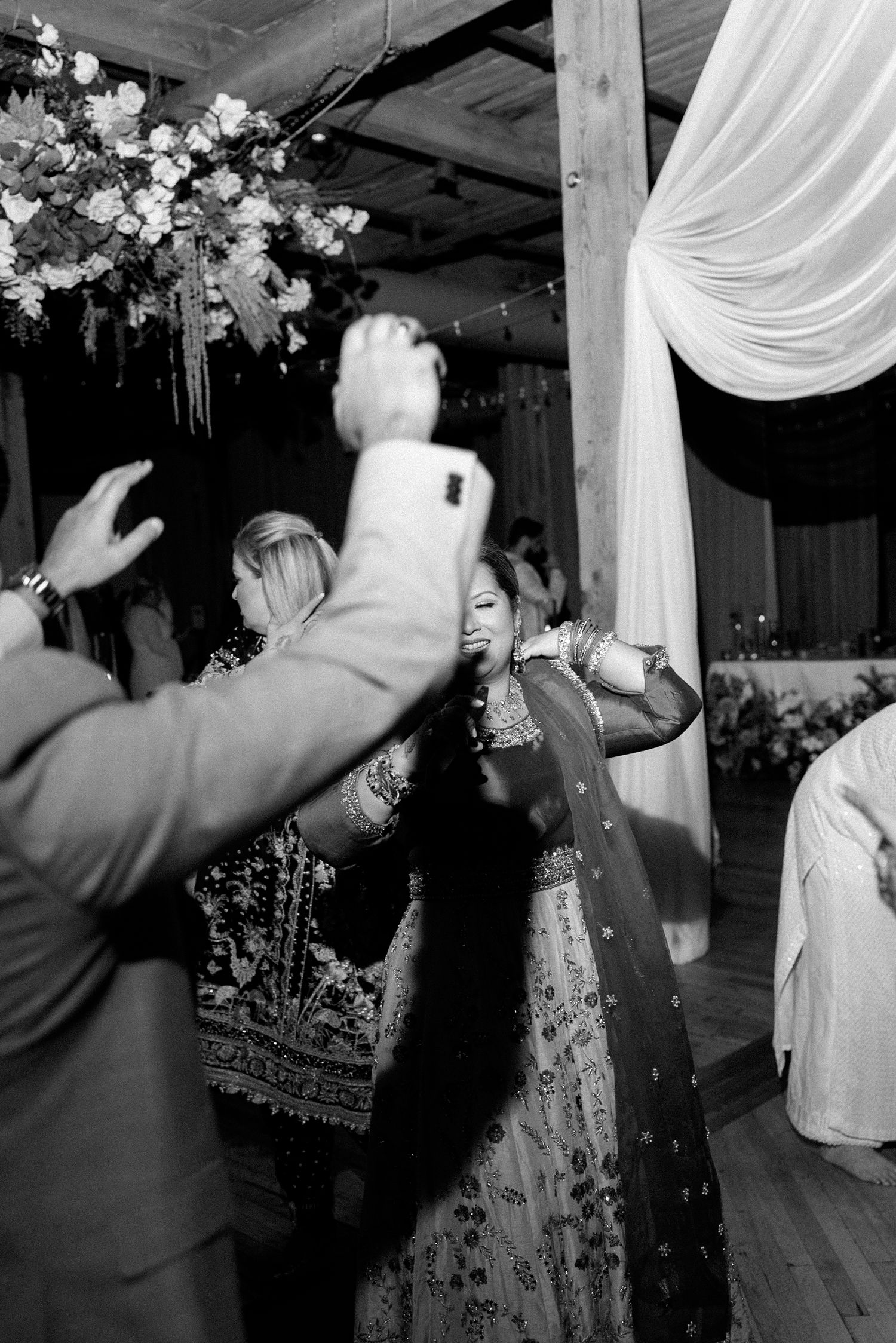 Black and white photo of guests dancing at an Indian wedding reception with floral decor and draped fabric.