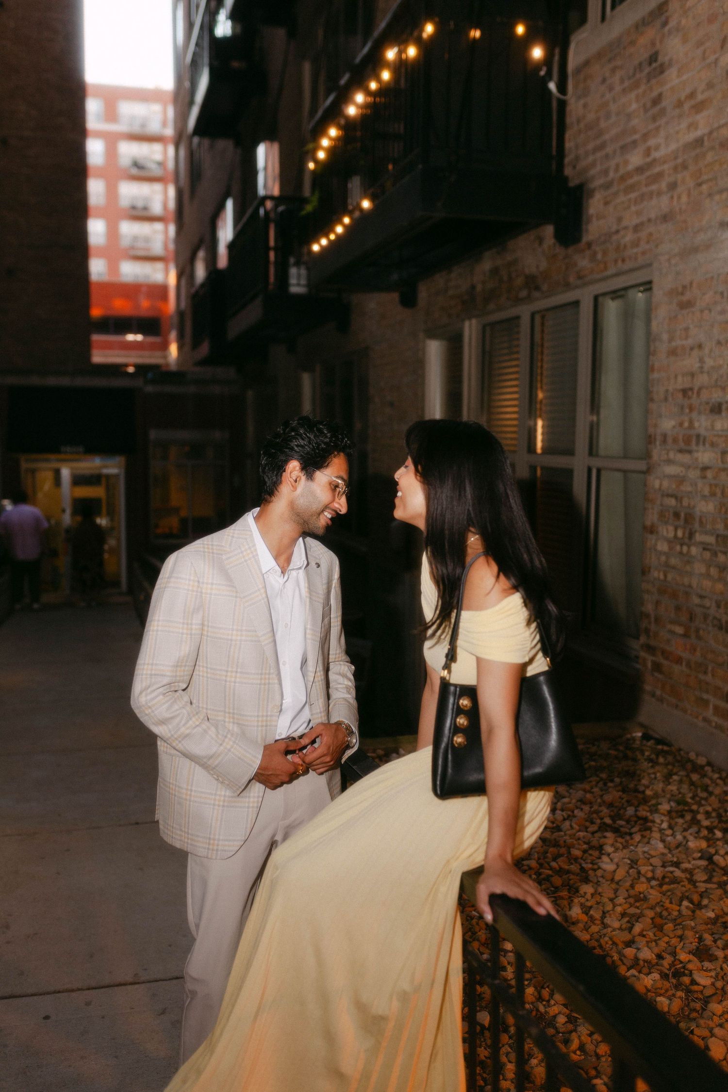 Couple in elegant attire sharing a romantic moment in a dimly lit urban alleyway with string lights above.
