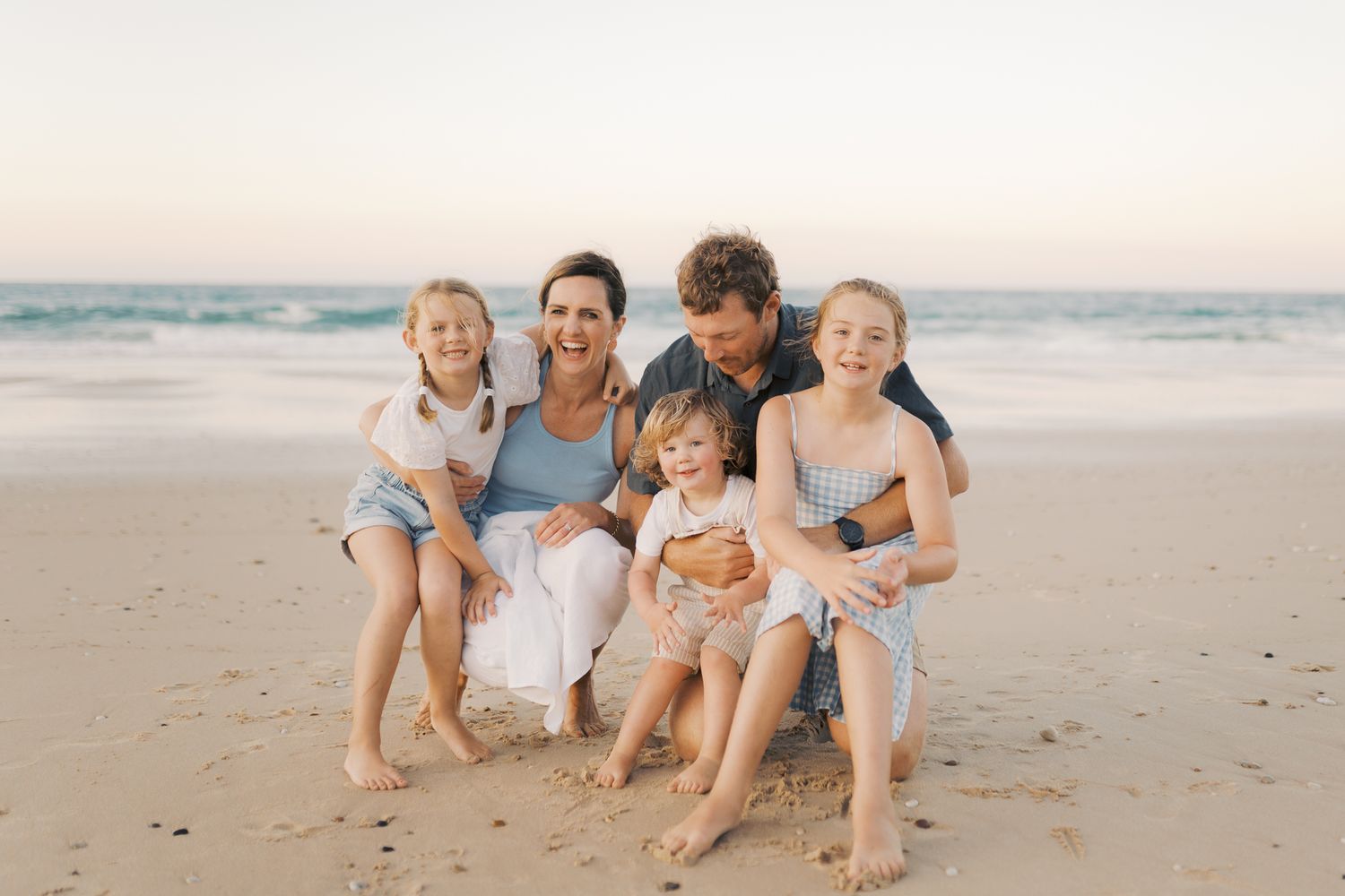 Family of five dressed in blue hues embracing while kneeling at the beach, Mum and children smiling at camera and Dad is looking at youngest