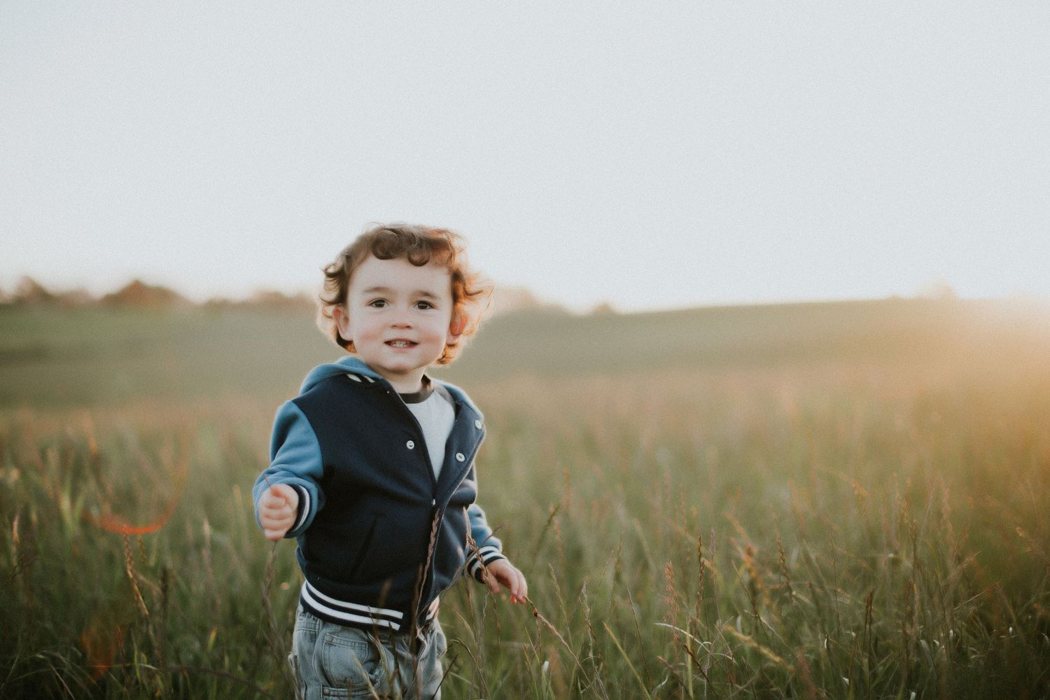 Toddler boy standing in field of tall green grass at sunset.